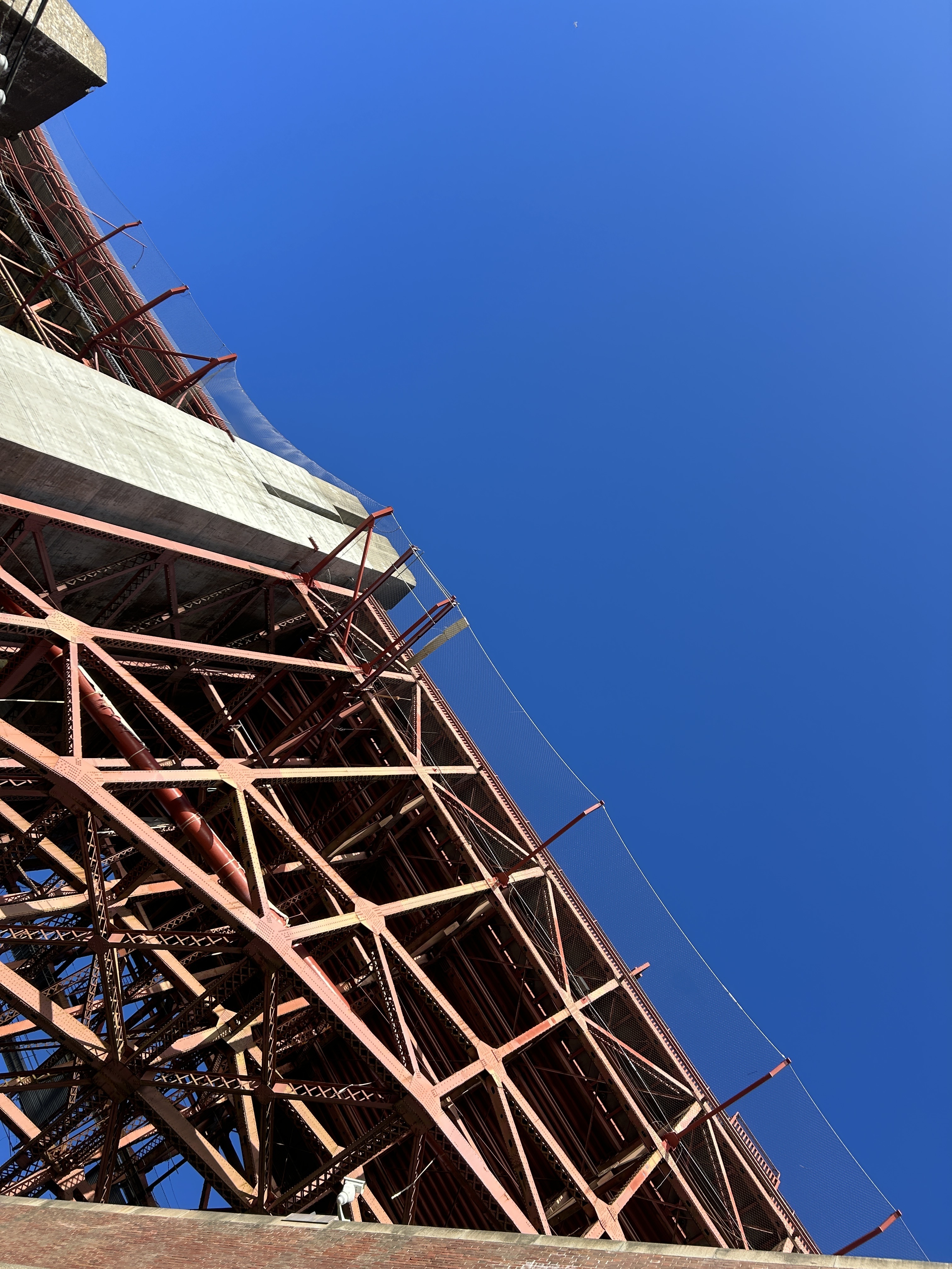 The image shows the underside of a red steel bridge structure against a clear blue sky. Diagonal metal beams and nets are visible, with some concrete elements.