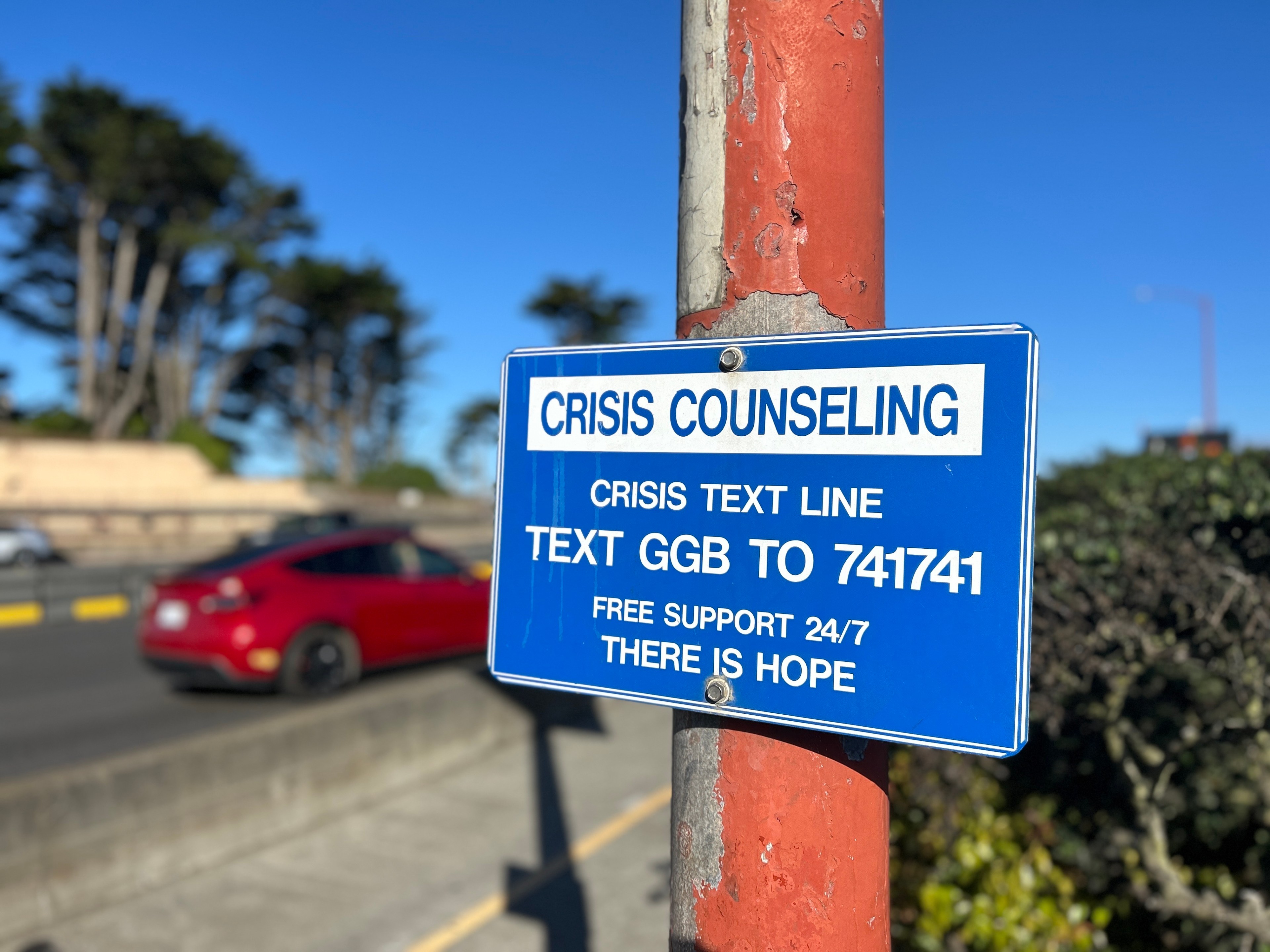 A blue sign on a red pole offers crisis counseling, instructing to text &quot;GGB to 741741&quot; for free 24/7 support. A red car and trees are visible in the background.