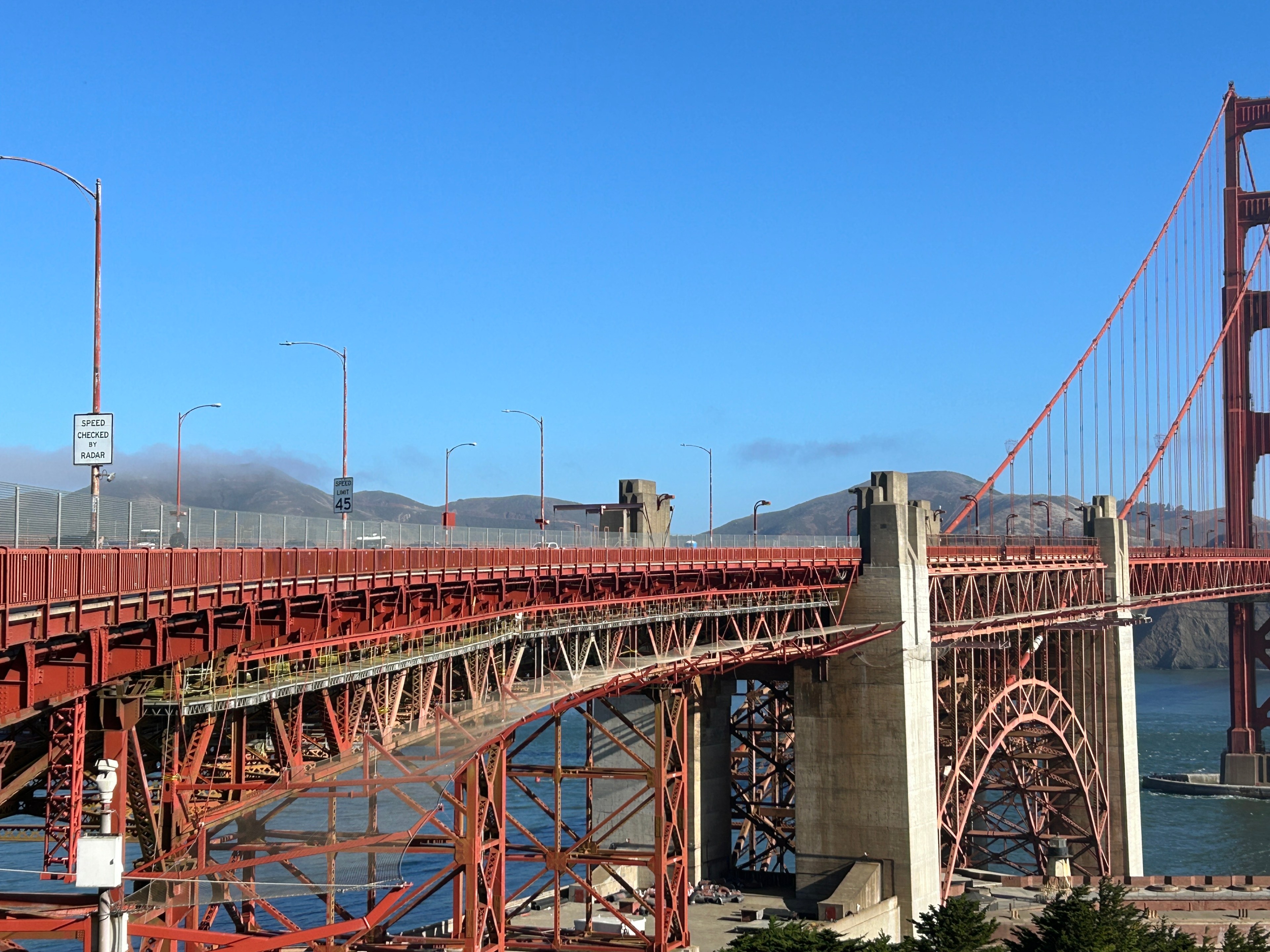 The image displays the Golden Gate Bridge, an iconic red suspension bridge, with clear skies above and hills in the background. A &quot;Speed Checked by Radar&quot; sign is visible.