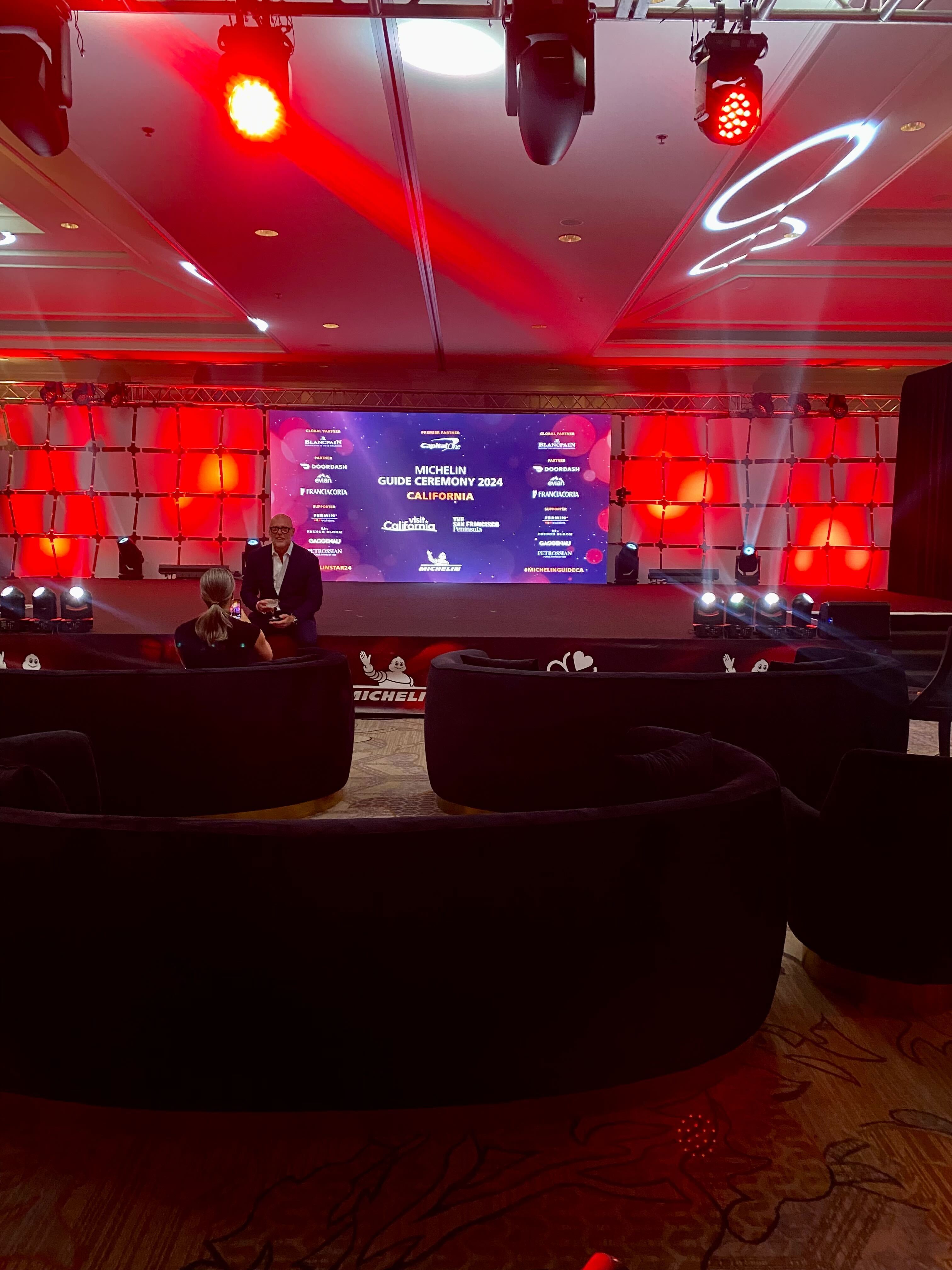 A photo of a low stage lit with red lights. A screen reads &quot;Michelin Guide Ceremoney 2024 California.&quot;