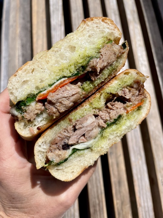 A hand holds a sandwich cut in half, revealing layers of meat, spinach, roasted red peppers, and a green spread, all encased in crusty bread.