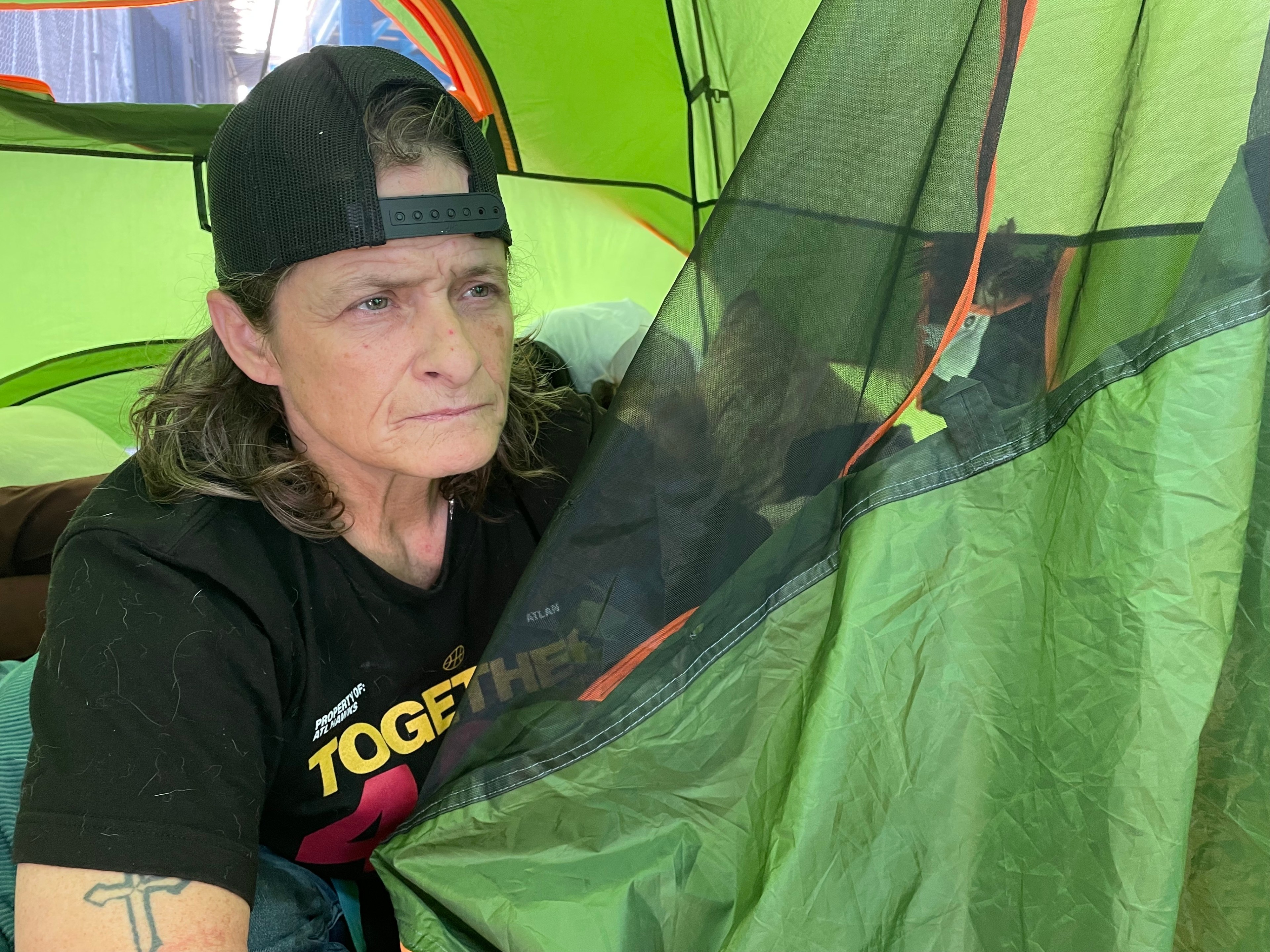 A person with long hair wearing a backwards cap sits inside a green tent, holding the tent flap. The person has a thoughtful expression and a cross tattoo on their arm.