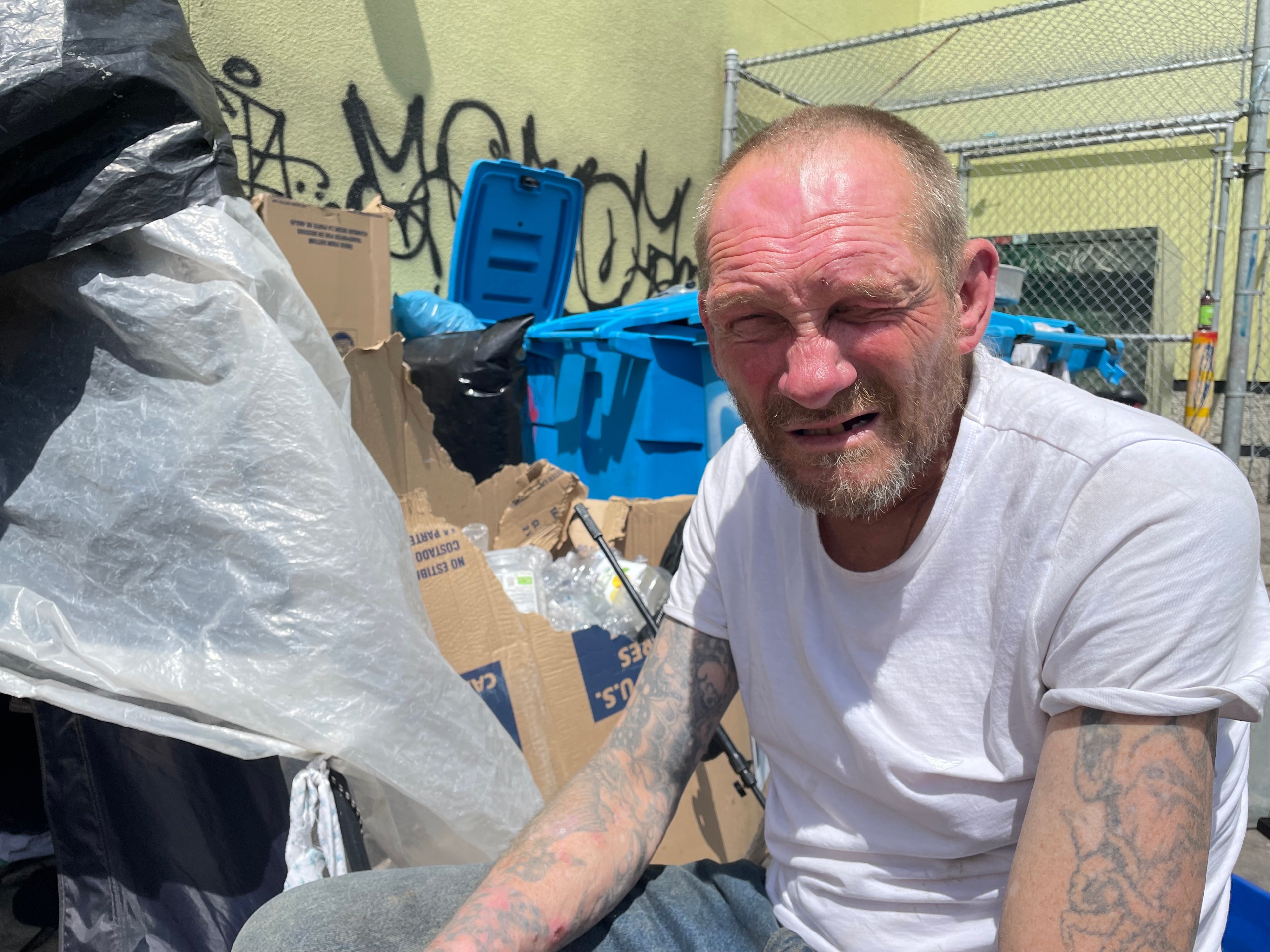 A man in a white T-shirt sits outdoors near cardboard boxes and blue bins. The area has graffiti and a metal fence in the background. His expression seems tense.