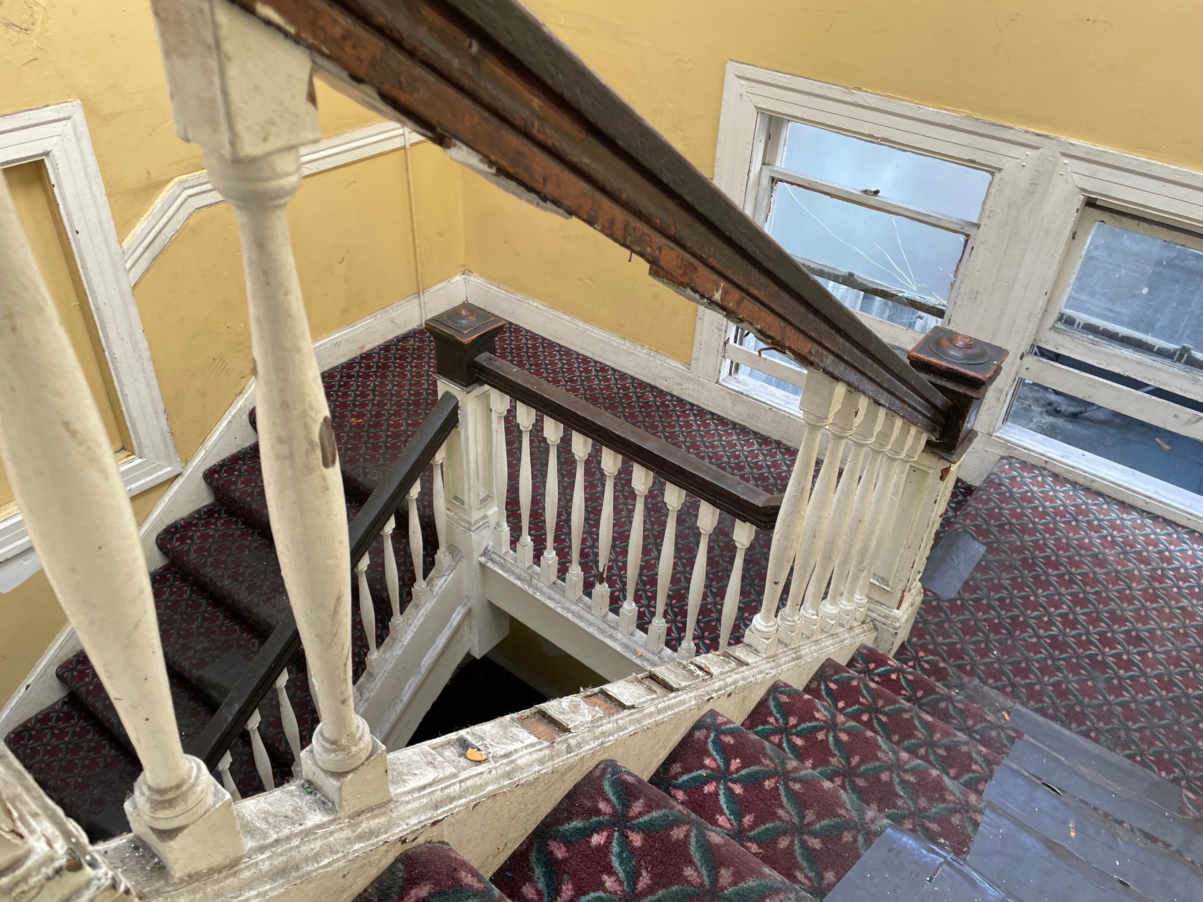 A worn, carpeted staircase with red and white geometric patterns descends between yellow walls. White railings frame the stairs, and large, old windows are visible.