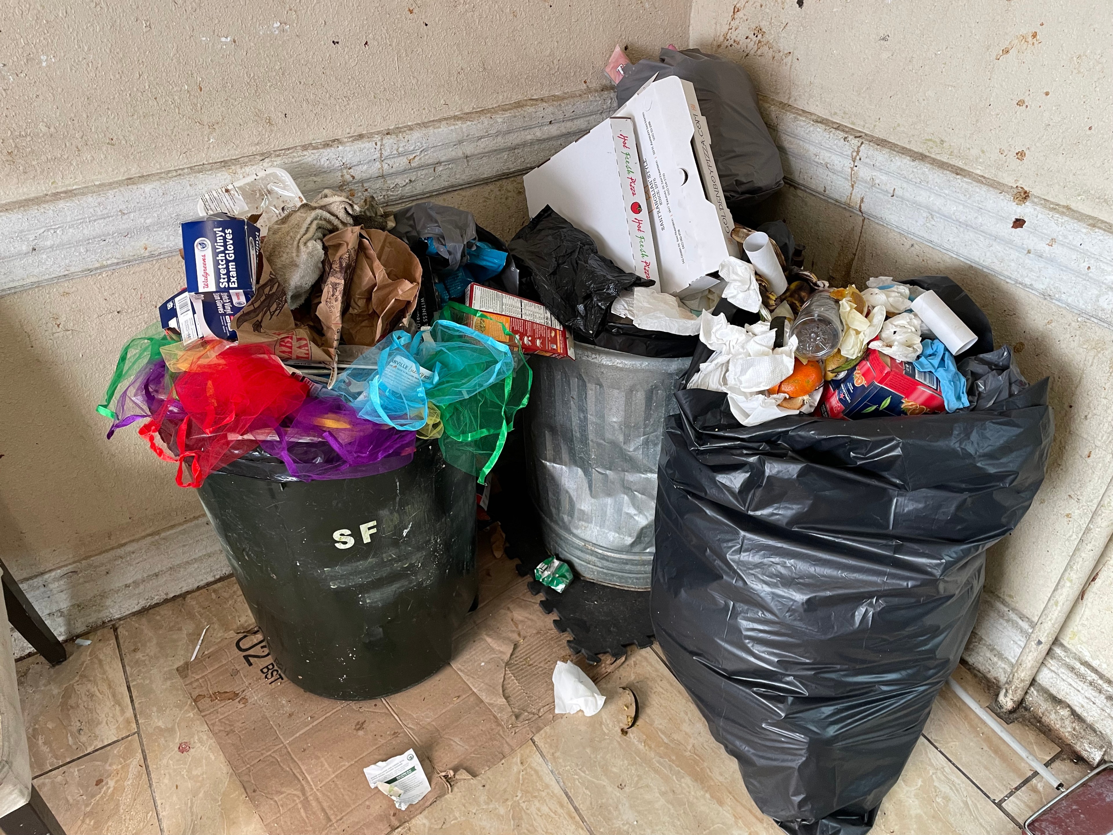 Three overflowing trash cans with various items like cardboard, packaging, and colorful nettings stand against a dingy wall, with debris scattered on the floor.