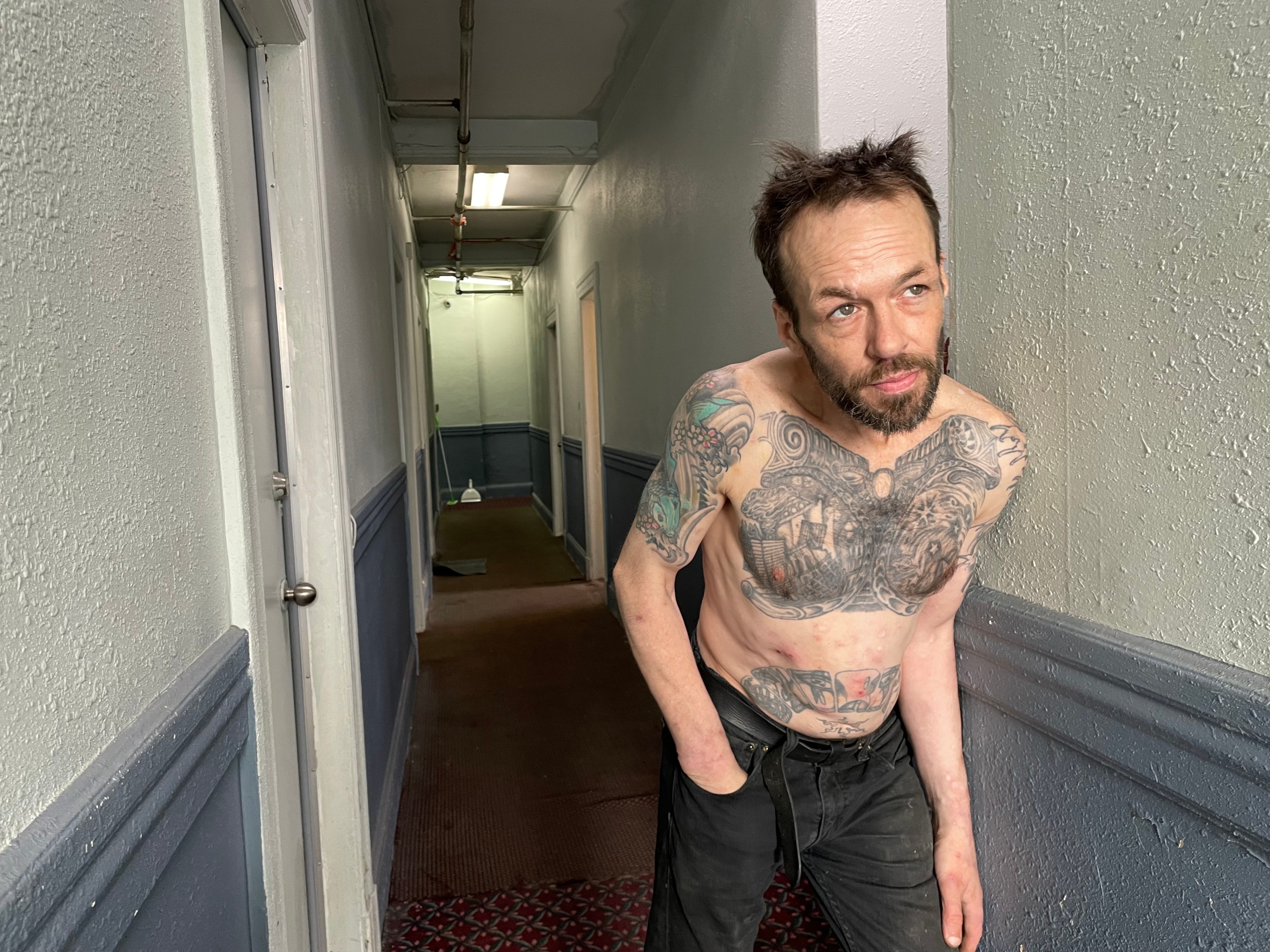 A shirtless, tattooed man with his left hand in his pocket leans against a wall in a narrow, dimly lit hallway, looking contemplative.