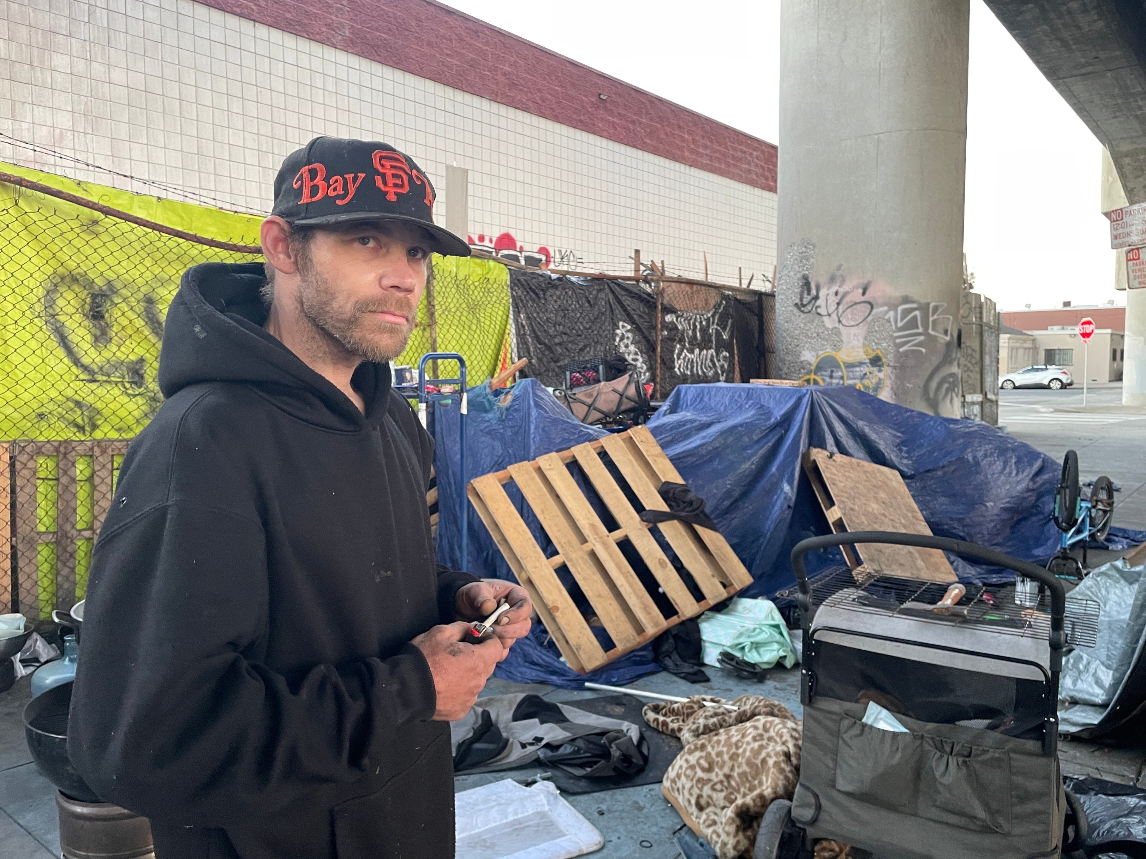 A man in a hoodie and cap stands in front of an improvised encampment under a bridge, surrounded by tarps, wooden pallets, and various belongings, with graffiti visible nearby.