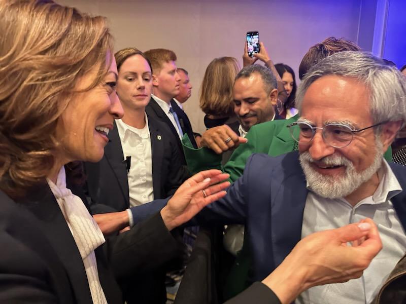 A woman in a suit smiles and greets an older man with glasses and a white beard. Several people, including security personnel, are gathered around them.