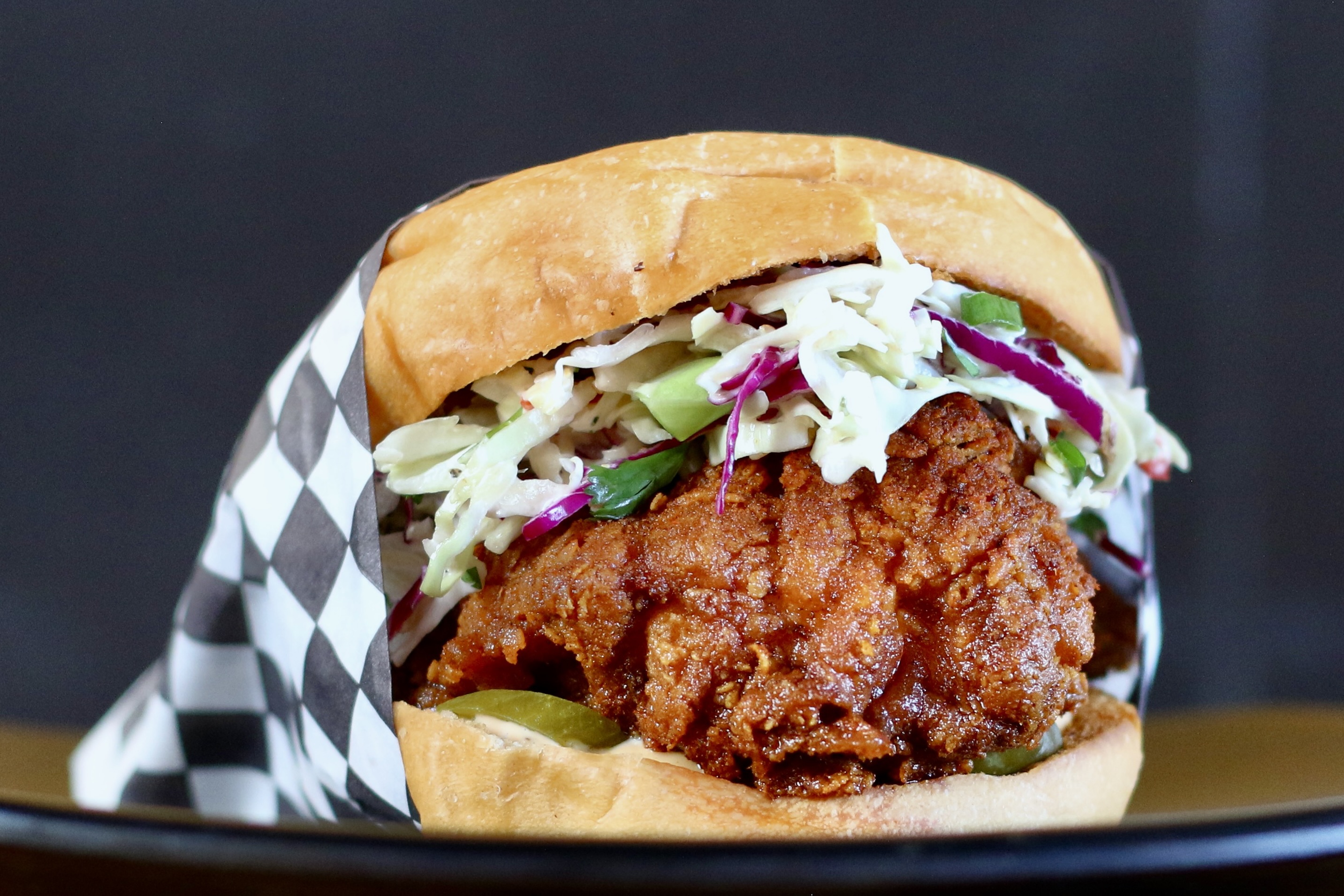 A crispy chicken sandwich with coleslaw and pickles is partially wrapped in black and white checkered paper, set against a dark background.