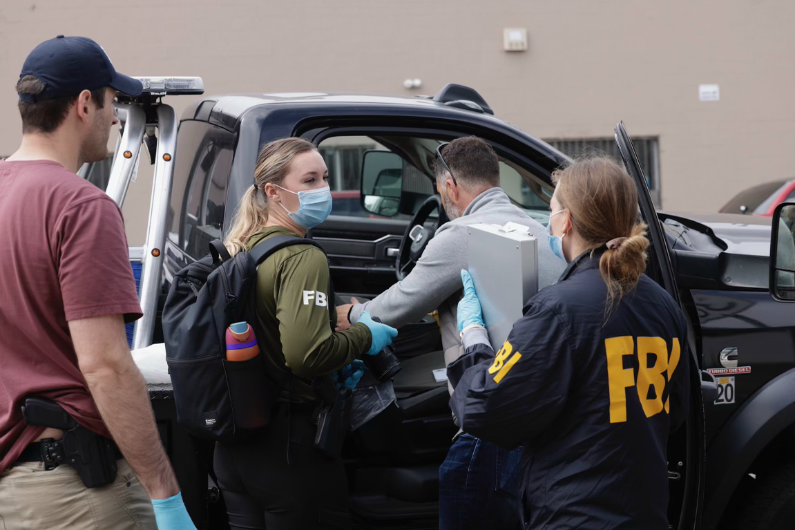 In this image, FBI agents, identified by jackets and a shirt, work around a pickup truck. They wear masks and gloves, handling a grey box, with another person nearby.