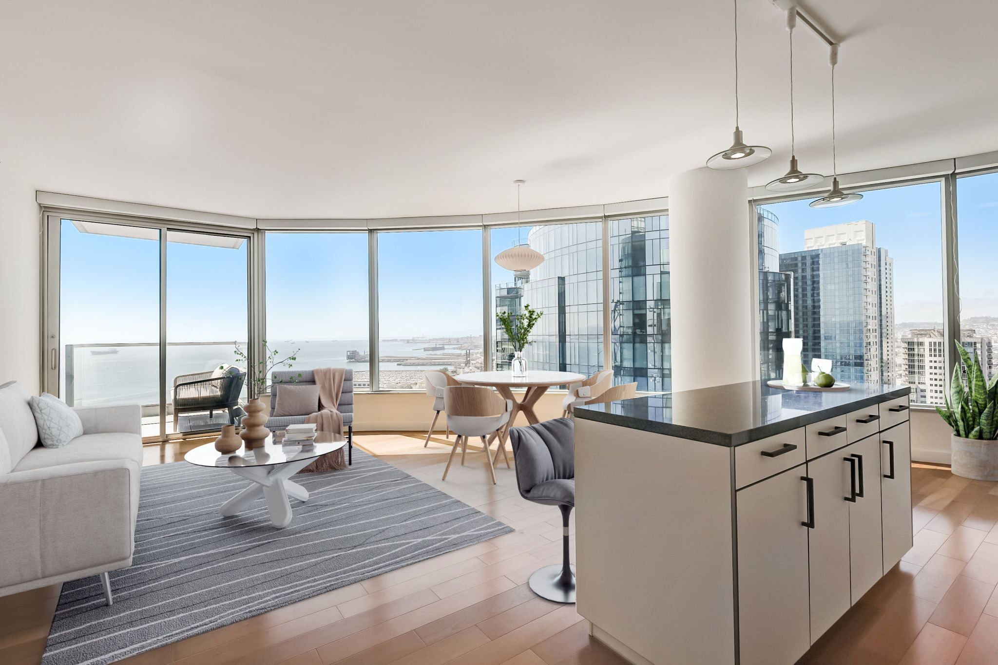 A modern, open-concept apartment has large windows with a city and water view, a cozy living area, a dining nook, and an island kitchen with sleek pendant lights.