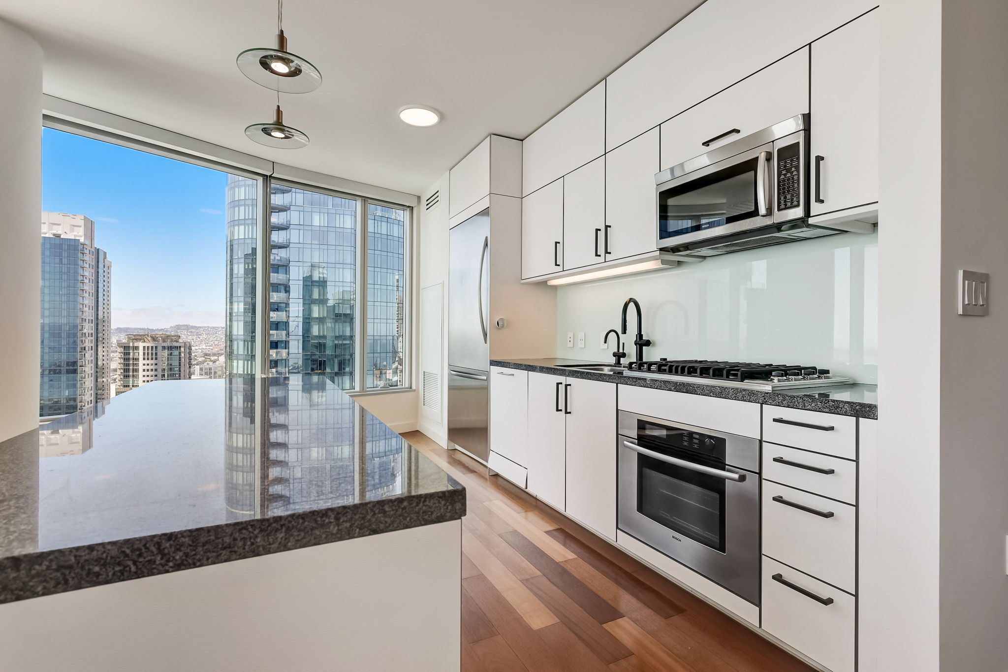 A modern kitchen with white cabinetry and stainless steel appliances overlooks a cityscape through large windows. Light wood flooring and sleek countertops complete the look.