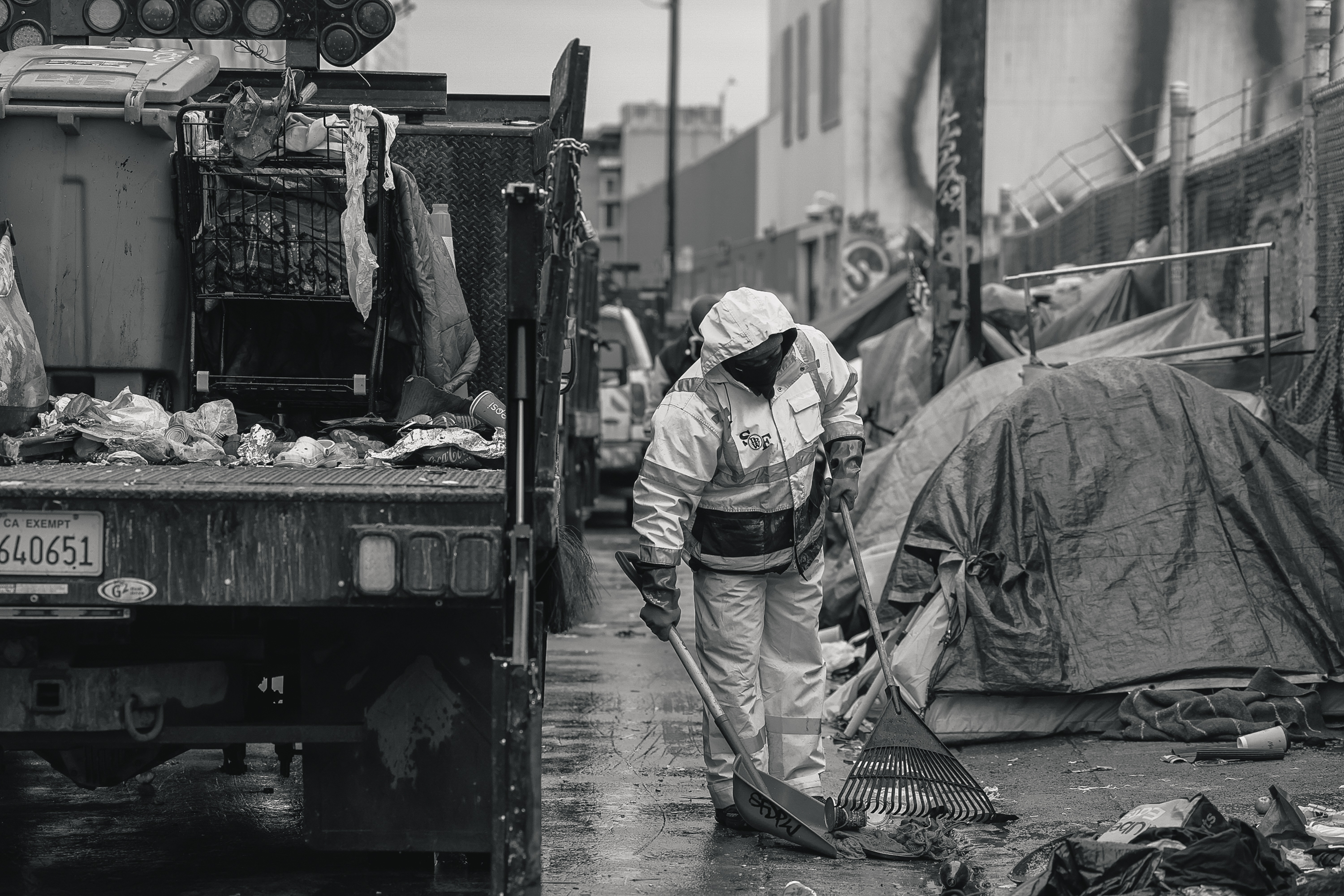 A person in protective clothing cleans a trash-strewn street with a rake near a truck loaded with garbage bags and gear, amidst makeshift tents and debris.