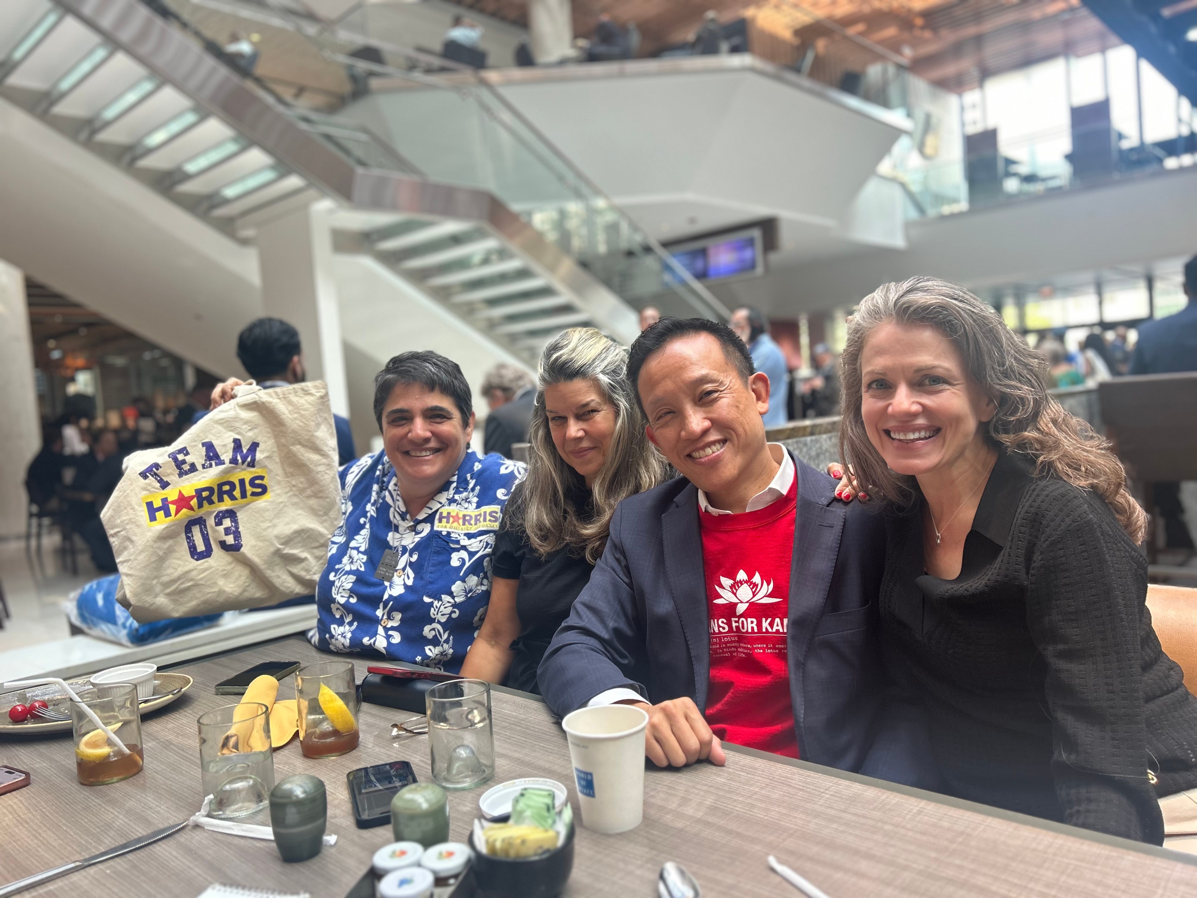 In a modern, indoor setting, four people sit at a table smiling, with one holding a "Team Harris 03" tote bag. Various drinks and snacks are on the table.