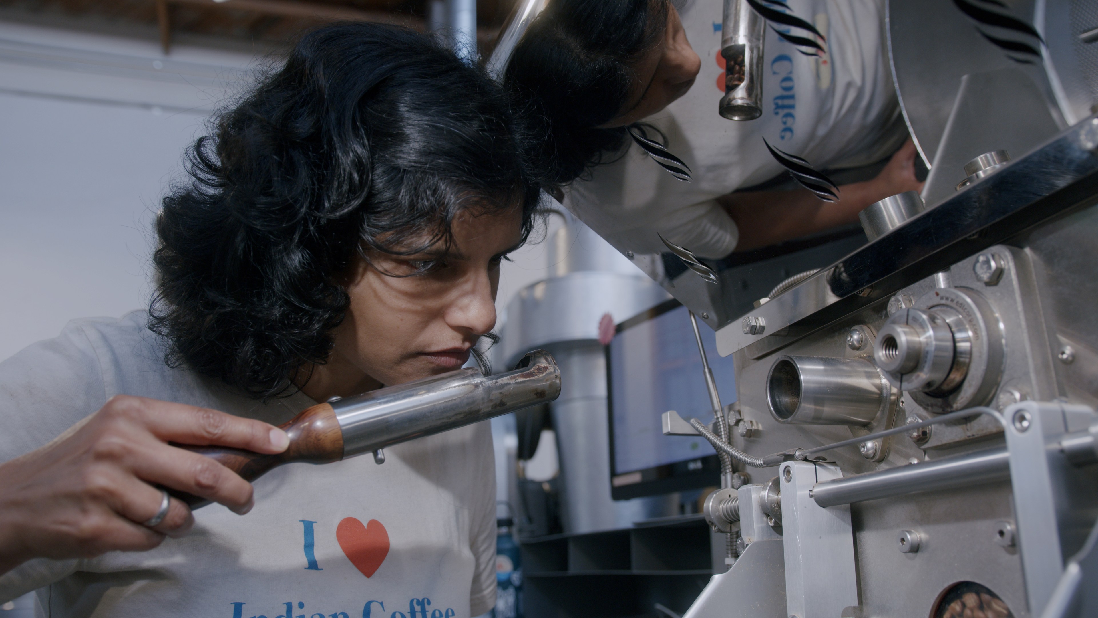 A person with curly hair inspects a steel machine, holding a tool. They wear a shirt with an &quot;I ♥ Indian Coffee&quot; text. The machine appears to be for coffee making.