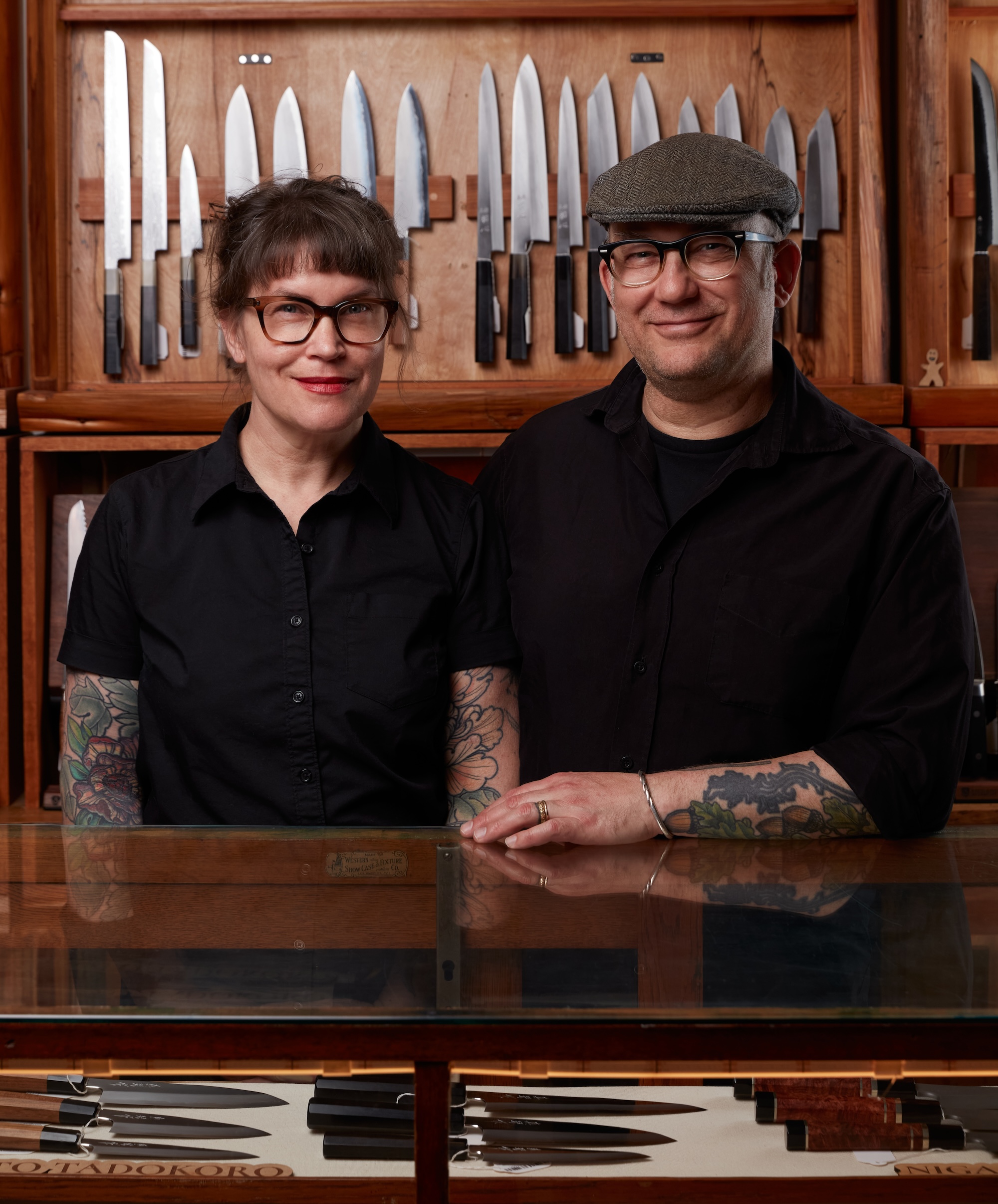 Two people, a women wearing glasses and a black shirt and a man wearing glasses and a hat also in a black shirt, stand behind a counter displaying knives.