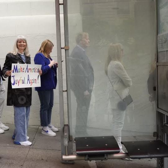 A woman in a black coat and blue jeans holds a sign saying &quot;Make America Joyful Again!&quot; while standing among a group of people, some looking at their phones, near a bus stop.