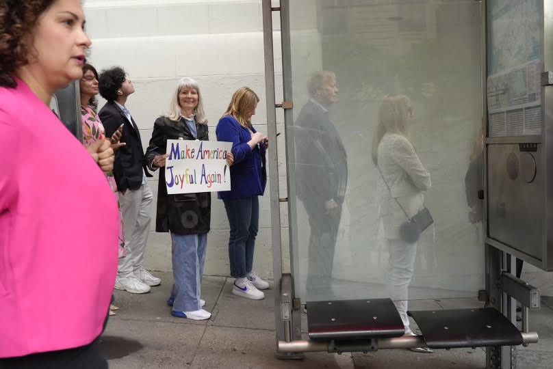 A woman in a black coat and blue jeans holds a sign saying &quot;Make America Joyful Again!&quot; while standing among a group of people, some looking at their phones, near a bus stop.