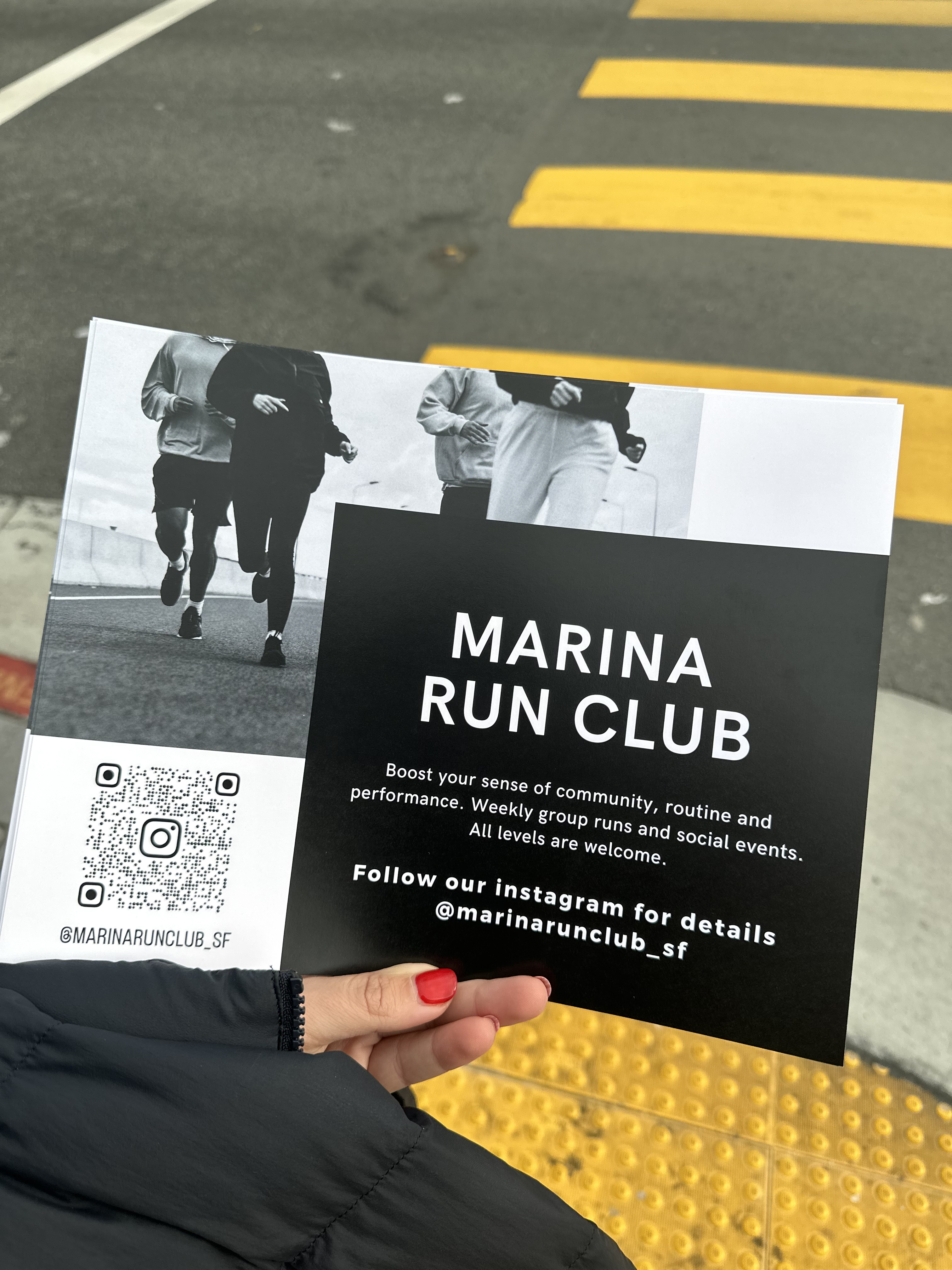 A person's hand with red nails holds a flyer for the Marina Run Club, which promotes community runs. The flyer includes a QR code and Instagram handle @marinarunclub_sf.