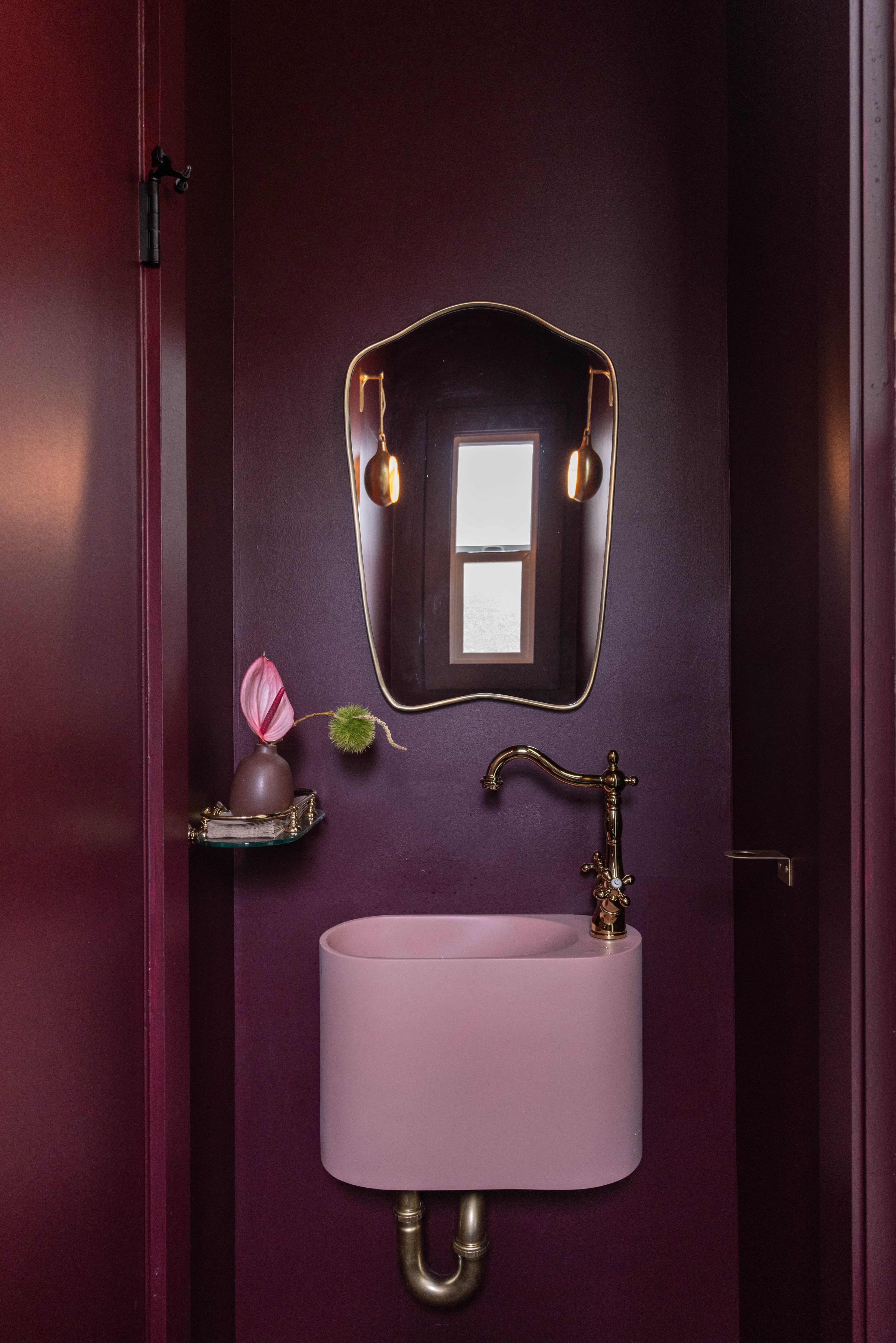 The image shows a small bathroom with dark purple walls, a pink sink, a brass faucet, a decorative mirror, a small shelf with a vase, and a window in the mirror's reflection.