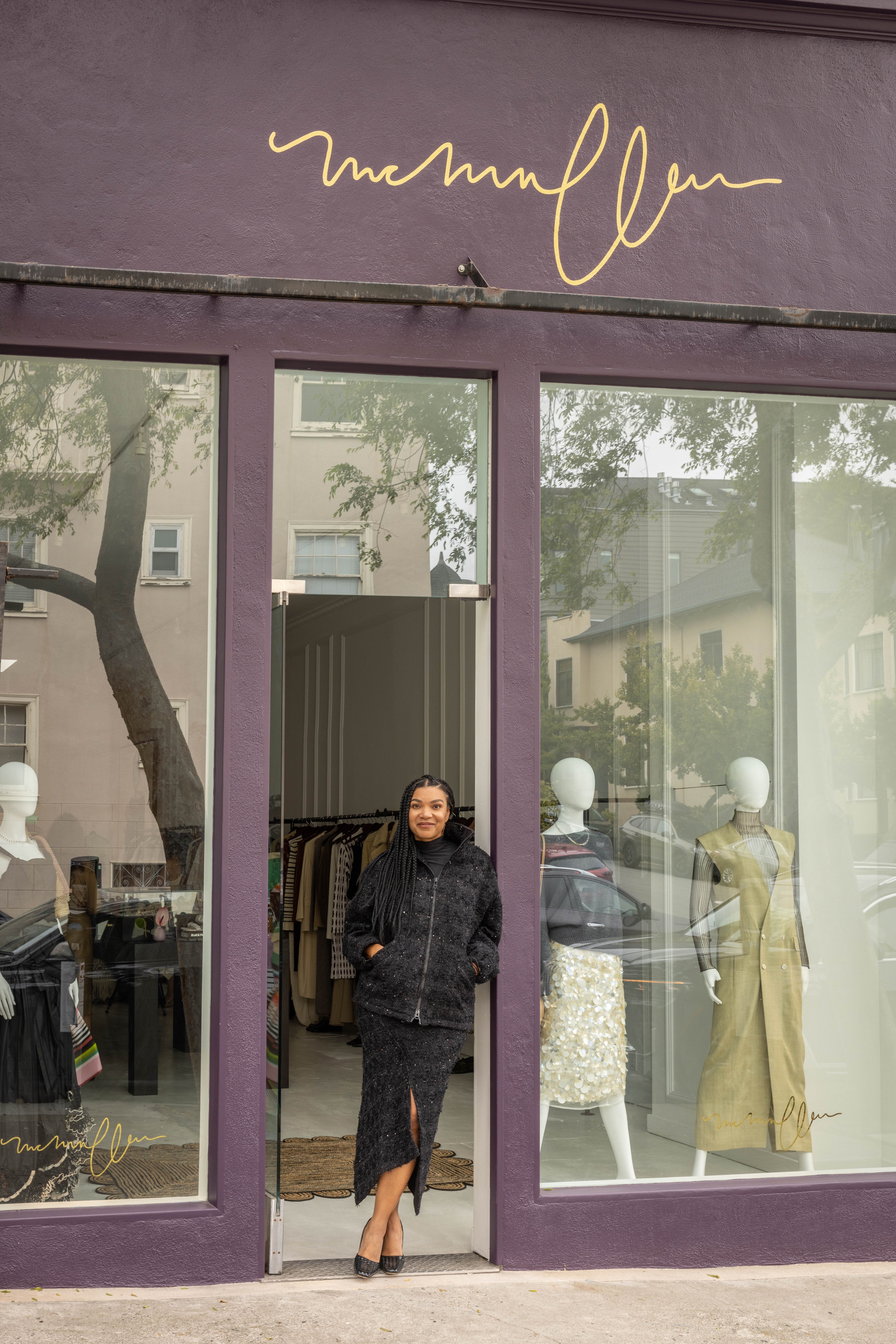 A smiling woman stands at the entrance of a fashion store with dark purple frames. Mannequins display outfits in the window, and the store's name is signed above.