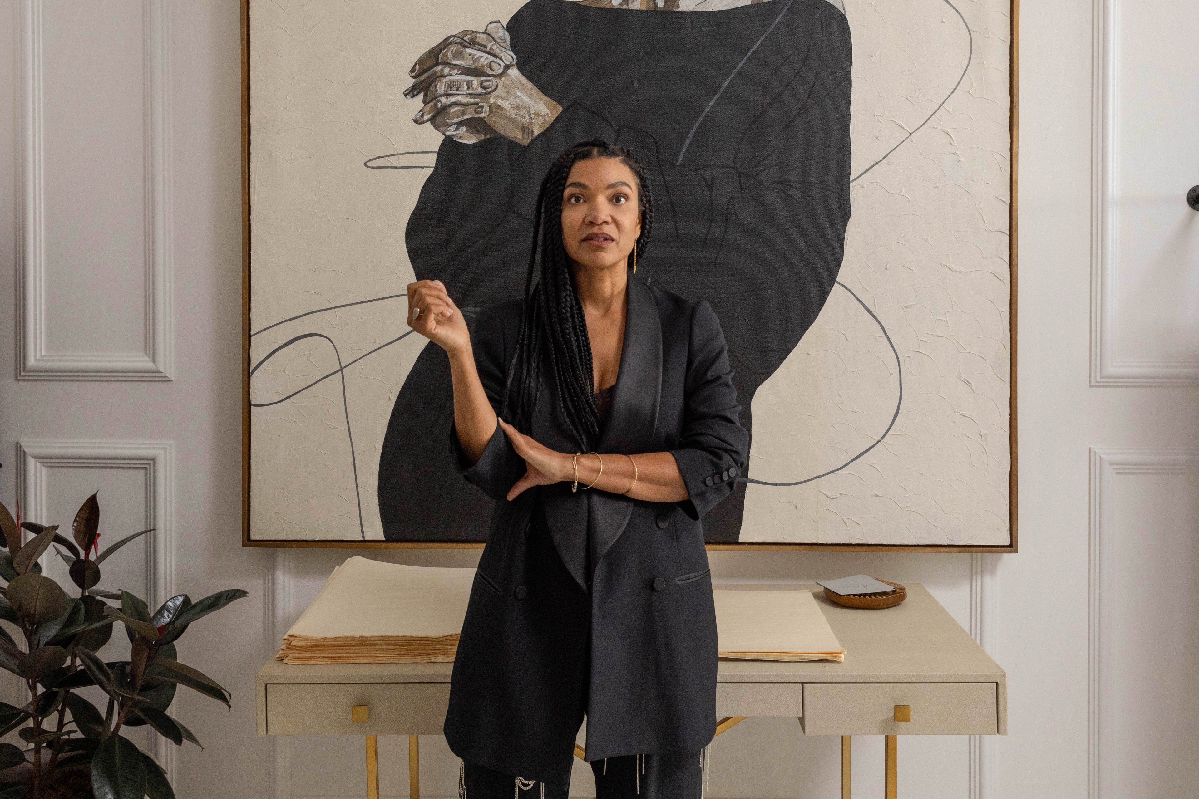 A woman stands in front of a large portrait wearing a black blazer and patterned black pants. A desk holds a stack of papers and a small plant.