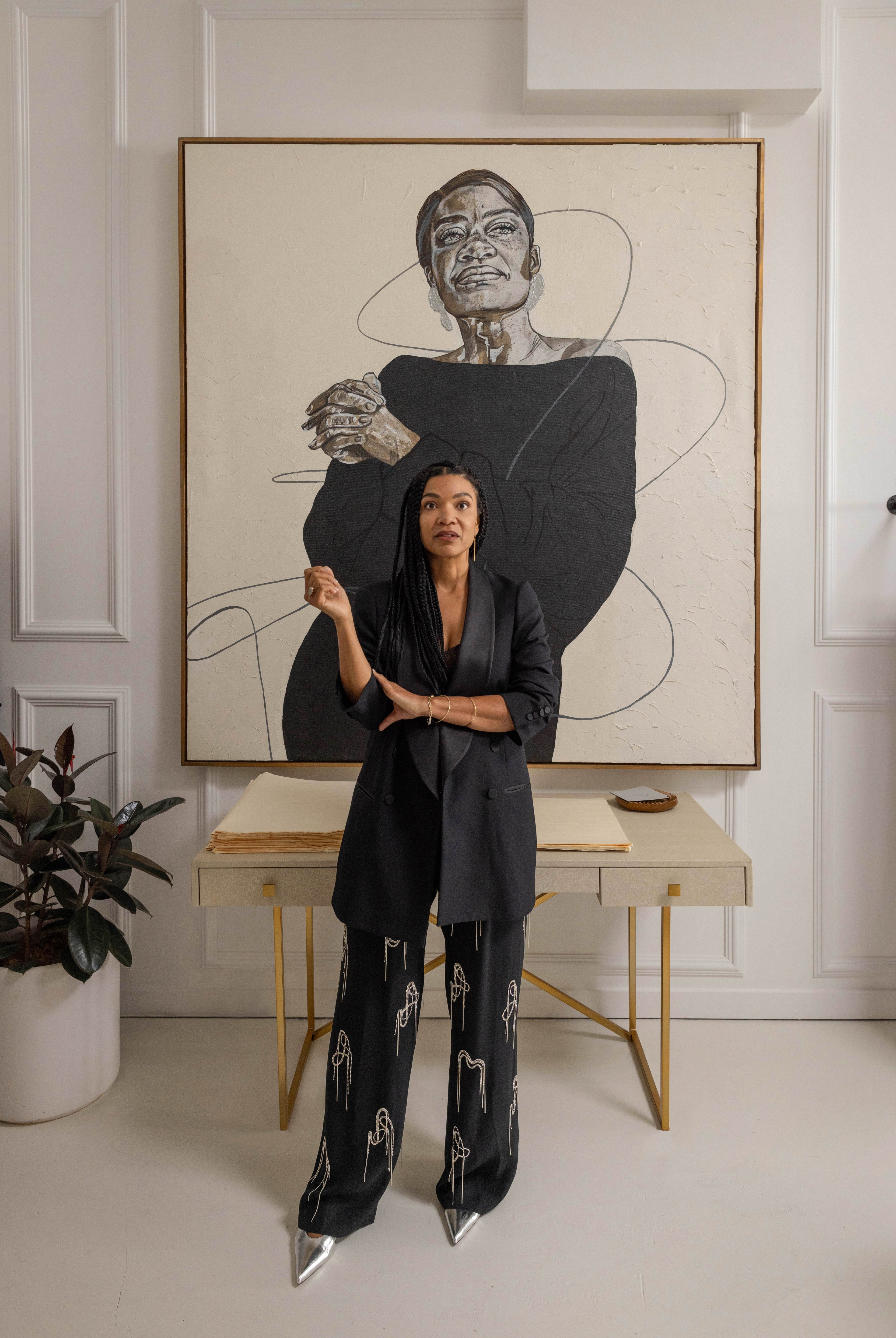 A woman stands in front of a large portrait wearing a black blazer and patterned black pants. A desk holds a stack of papers and a small plant.