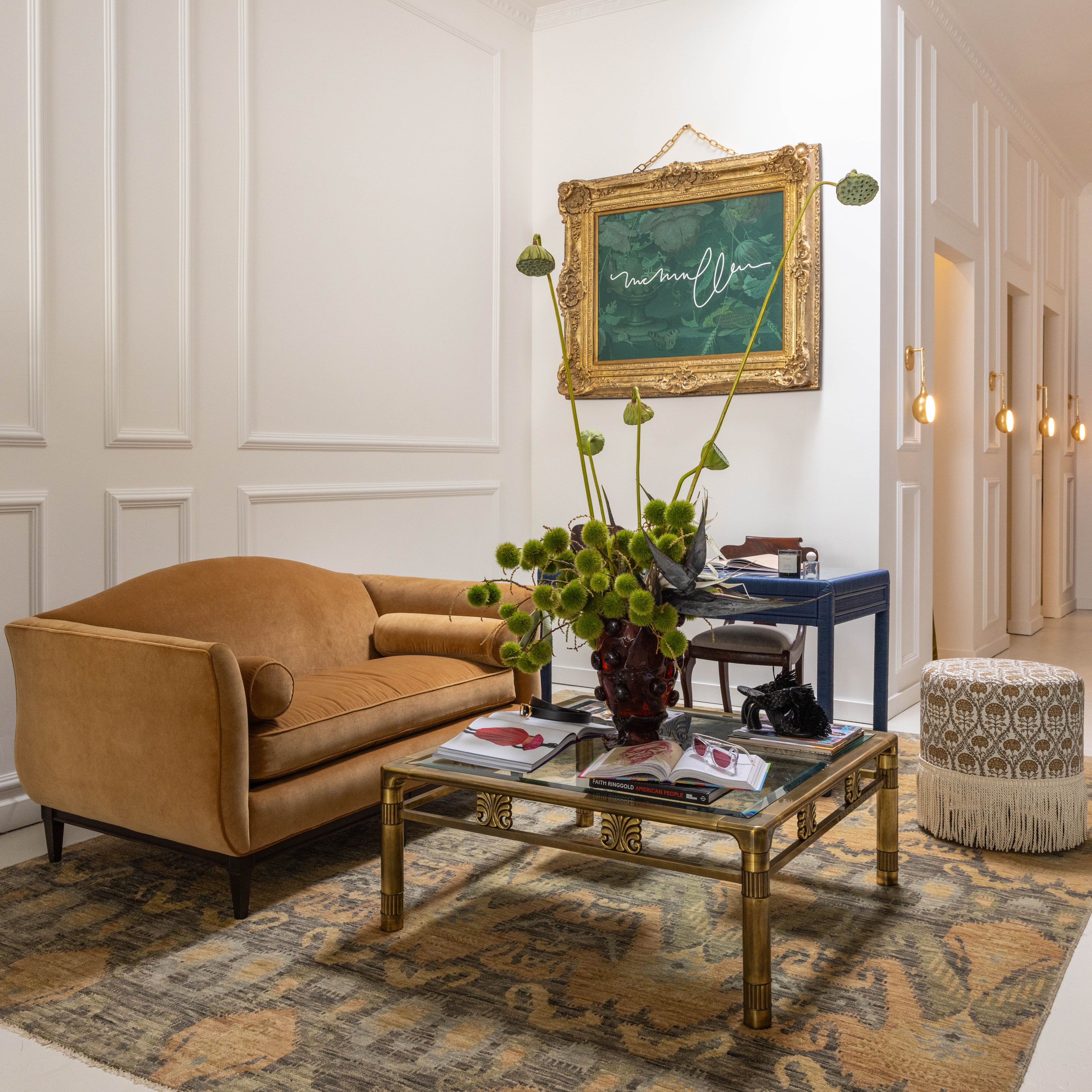 The image shows a room with a mustard-colored sofa, a glass coffee table with books, a large floral arrangement, a framed painting, and a pouf on a patterned rug.