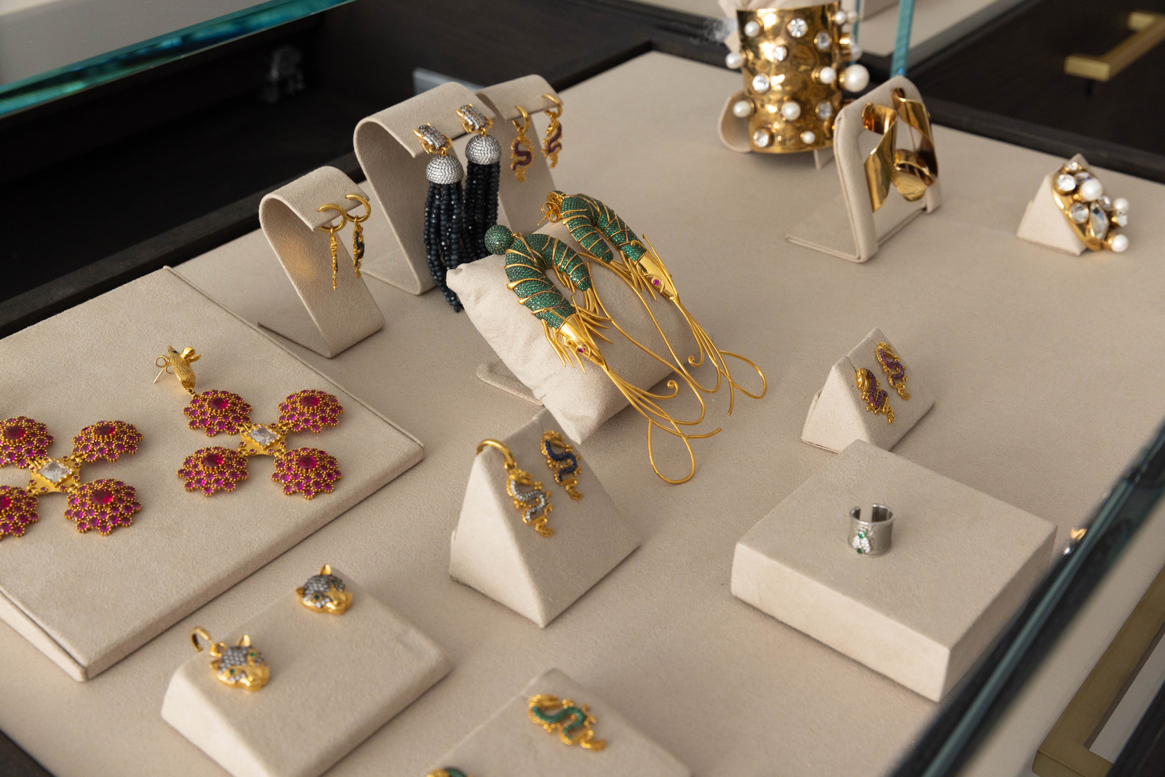 A jewelry display features ornate gold earrings, rings, and bracelets adorned with colorful gemstones and pearls, all arranged on beige stands.