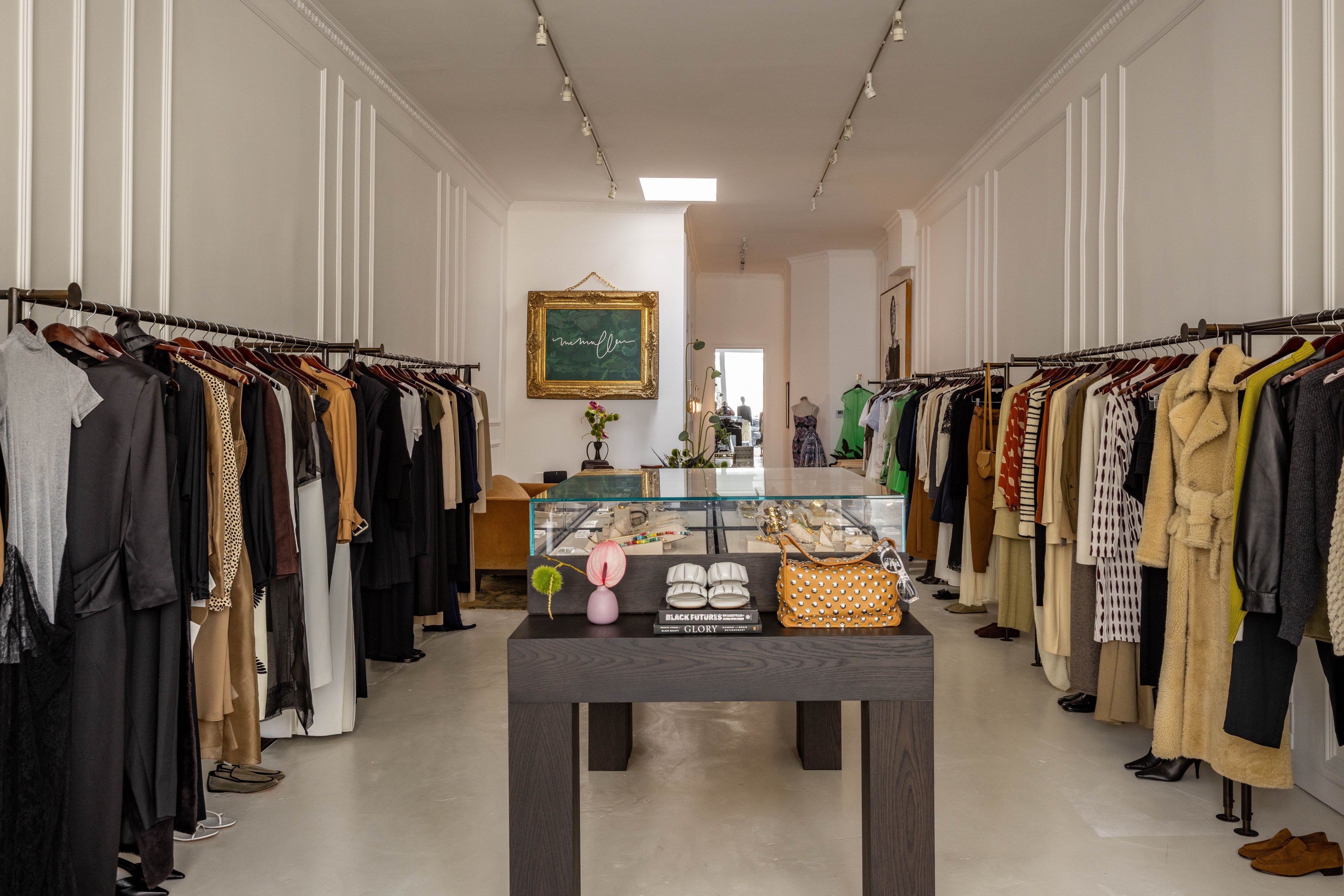 The image shows a boutique with clothing racks on both sides, a central display table with accessories, and a well-lit, elegant interior.