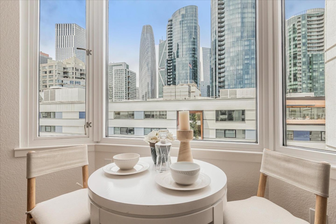 A small round table with two chairs is set by a corner window, offering a view of modern city skyscrapers. The table has two bowls, glasses, and candles.