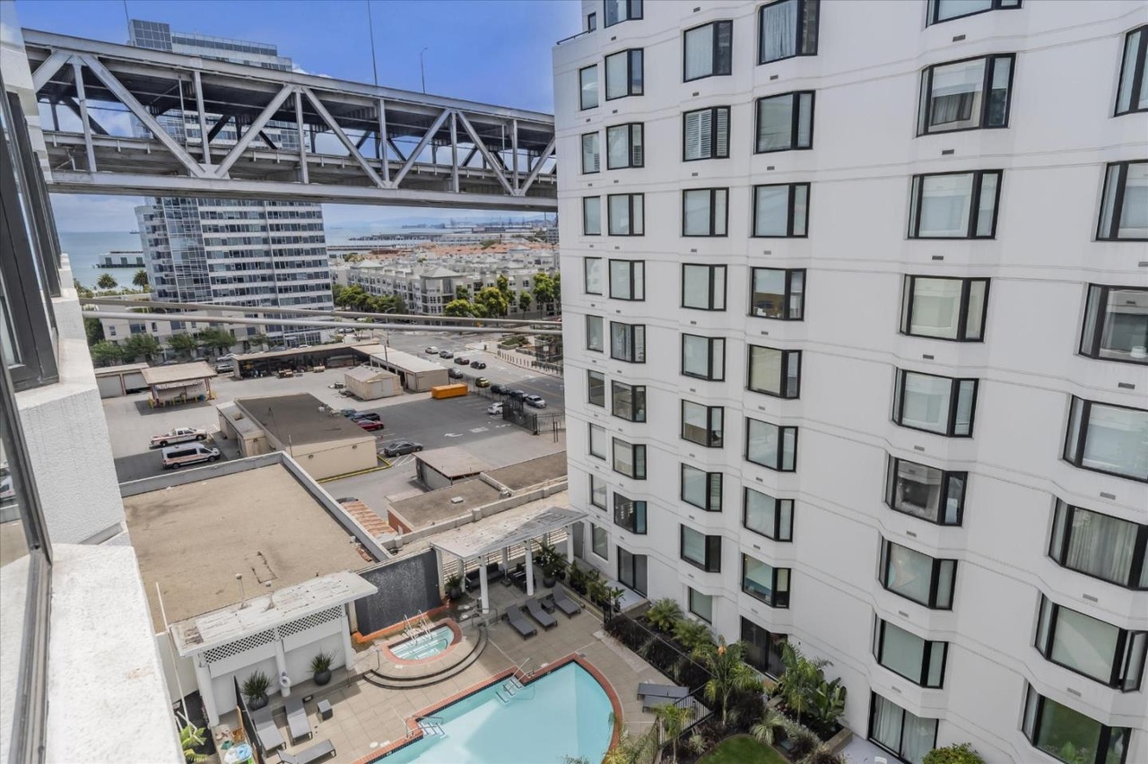 The image shows a modern urban scene with tall white buildings featuring numerous windows, a rooftop pool area below, and a bridge structure overhead. In the background, there is a partial view of a cityscape with water visible.