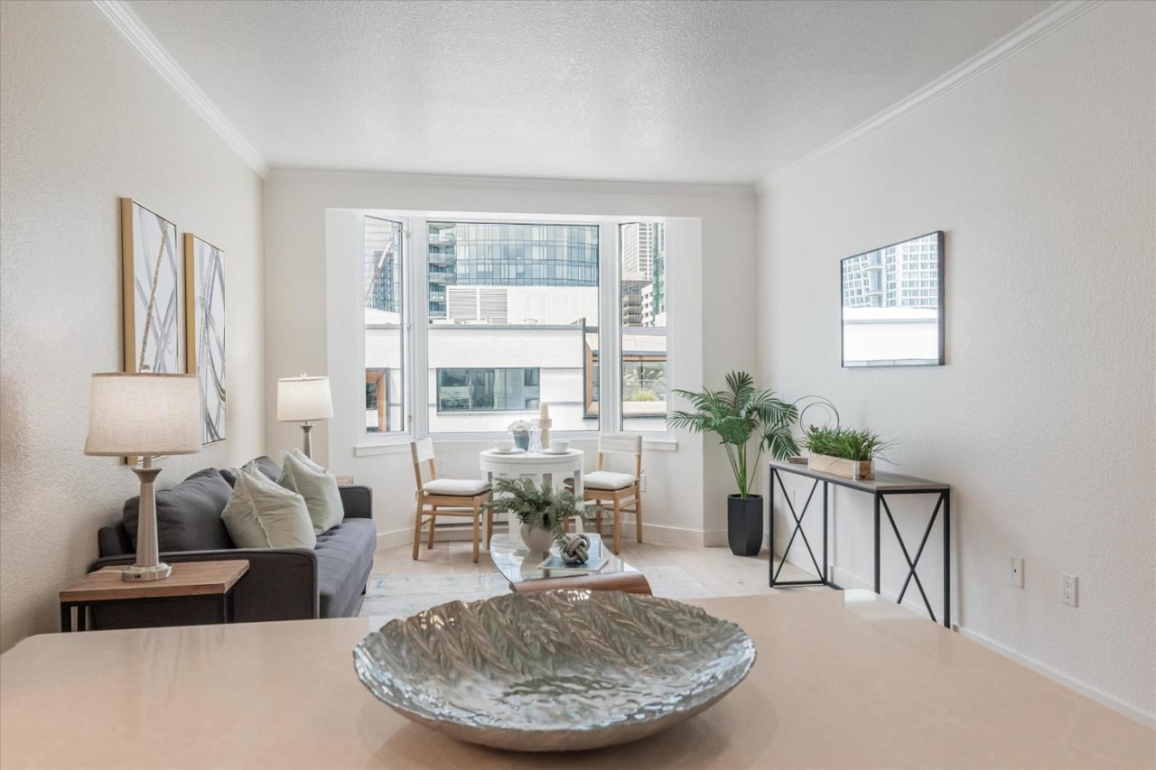 A cozy living room includes a gray sofa with cushions, two lamps, a coffee table, and wall art. By the window, there's a small dining table with chairs, and a console table with plants to the right.