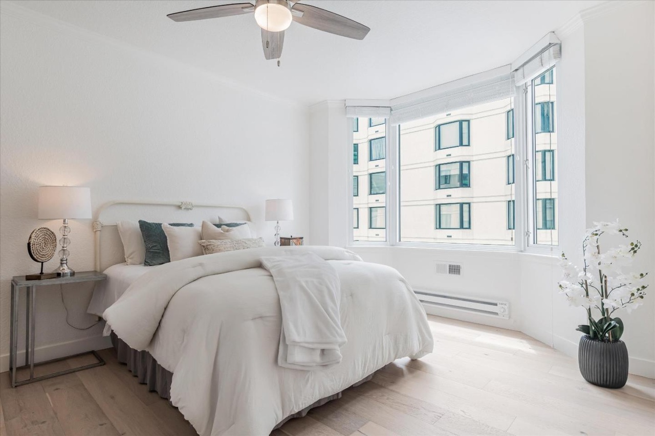 A bright bedroom features a neatly made bed with white linens, flanked by two bedside tables with lamps. There is a ceiling fan, large windows, and a potted plant.