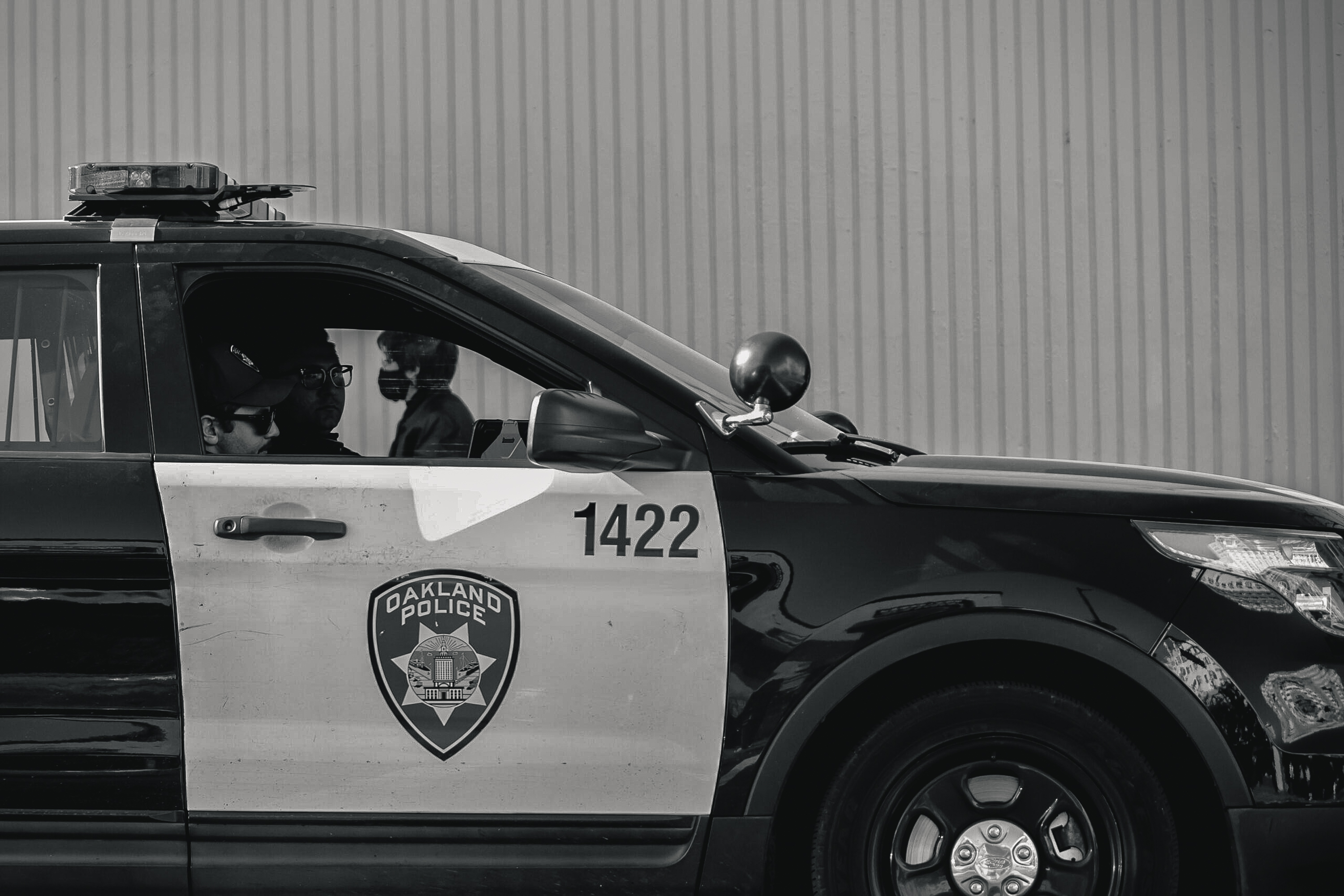 A black-and-white photo shows a parked Oakland Police car with two officers inside, one wearing glasses. The car has the number 1422 and a police emblem on the door.