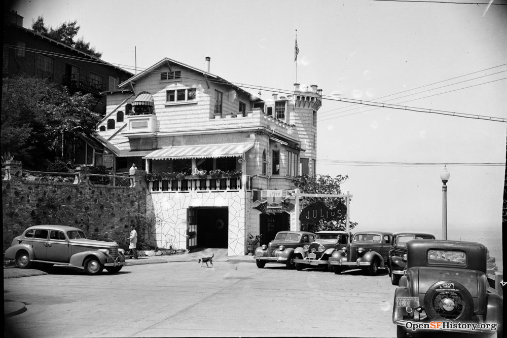 A historic building, with a castle-like tower and an American flag, stands above a garage. Several vintage cars and a pedestrian with a dog can also be seen.