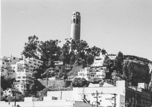 A tall tower stands atop a hill surrounded by trees and various buildings, with more structures and power lines visible in the foreground.