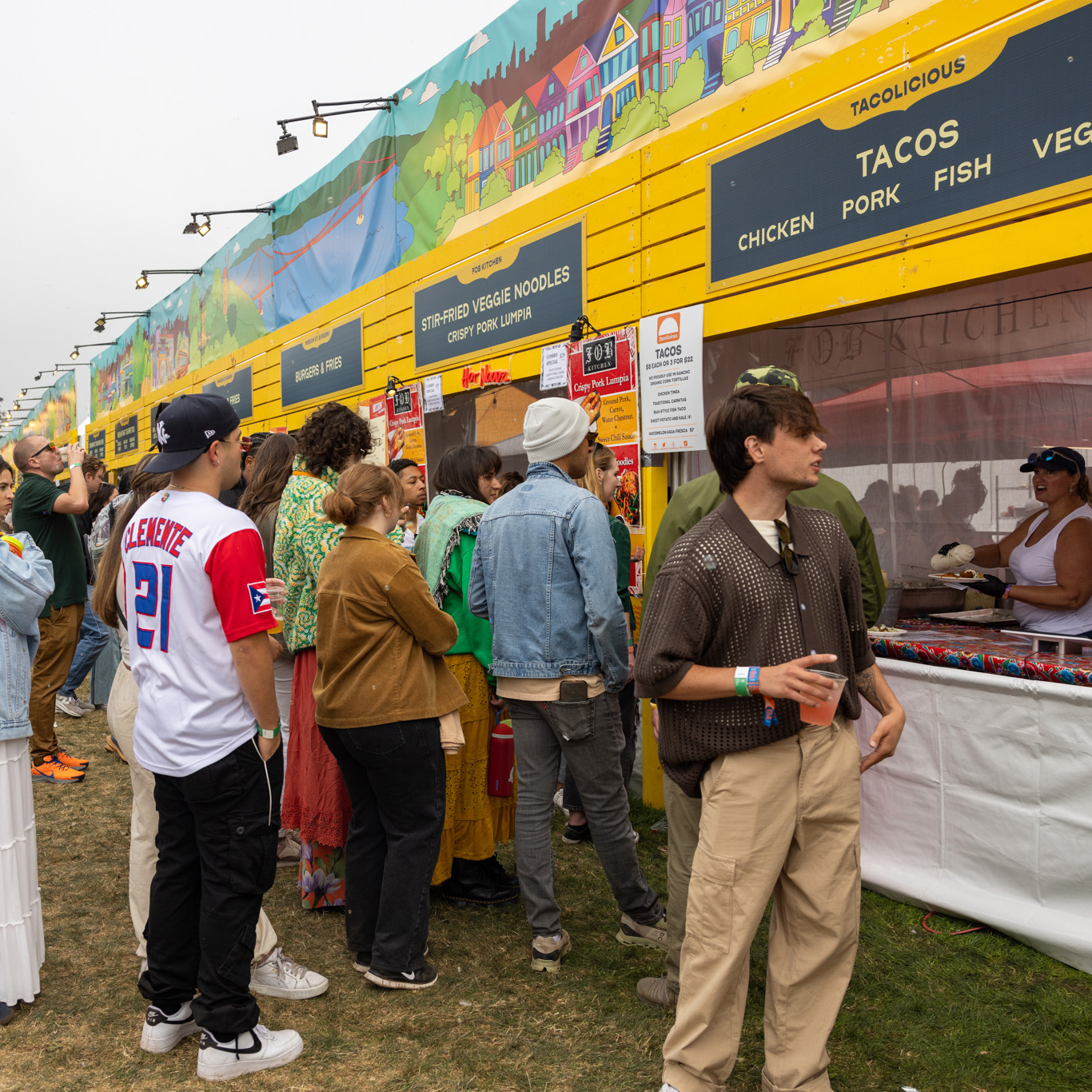 People are gathered in line at a vibrant food stall with a yellow facade offering tacos. The background features colorful mural art, and a person is looking at the menu.