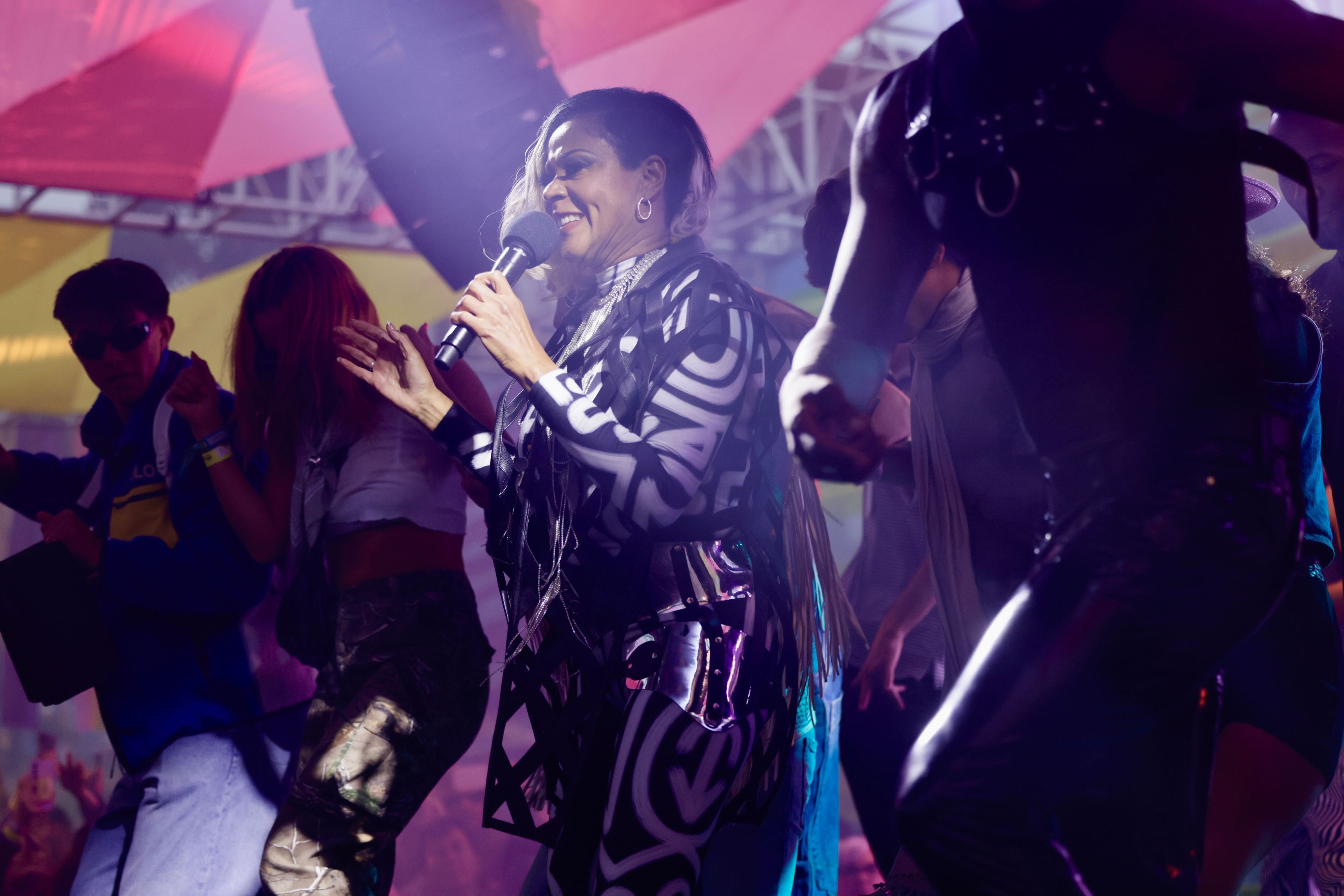 A woman in stylish black and white geometric patterned clothes sings into a microphone on stage, surrounded by energetic dancers under colorful lighting.