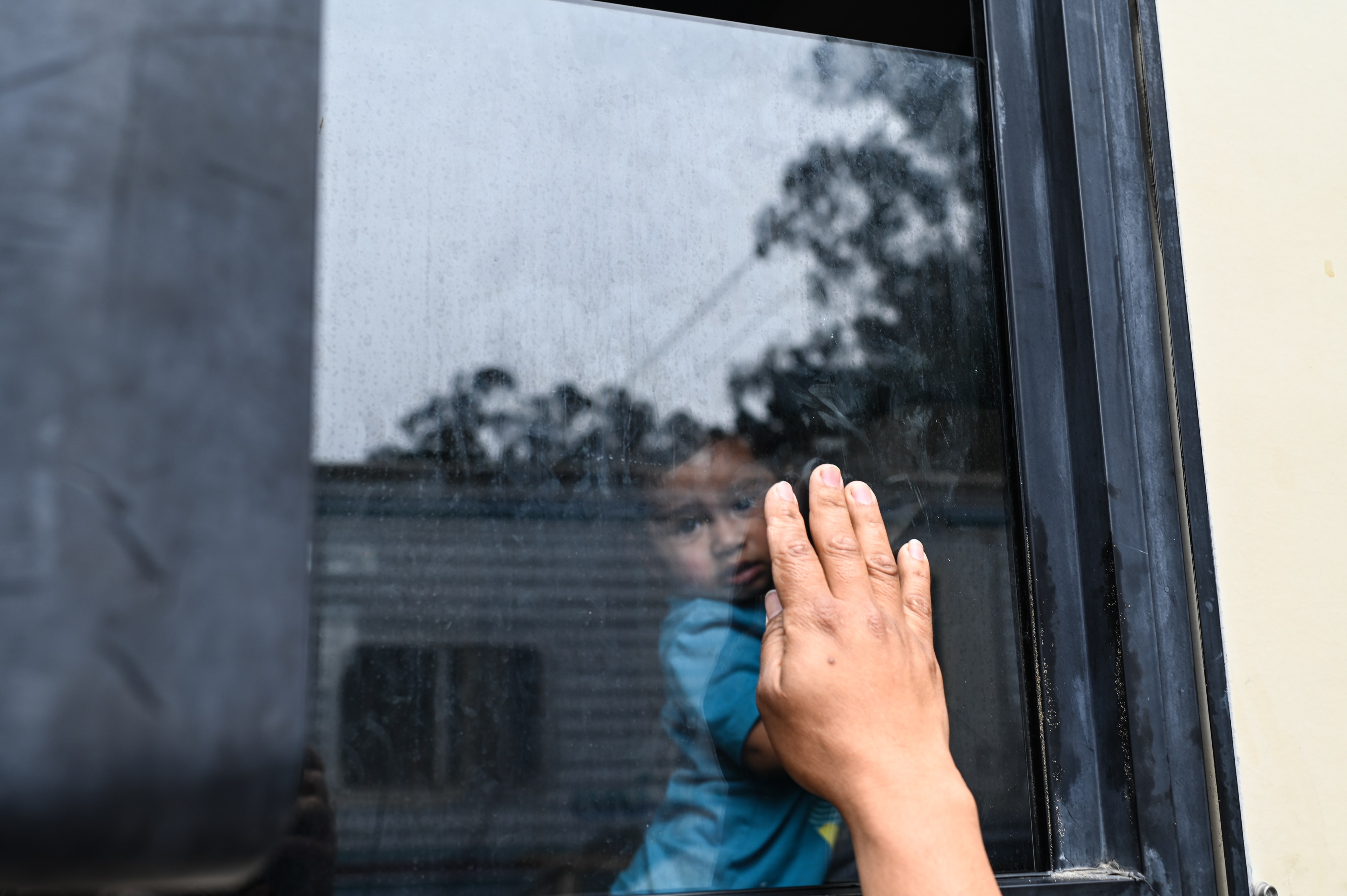 A hand touches a rain-speckled window, reflecting the image of a young child looking out, with blurred outdoor scenery in the background.