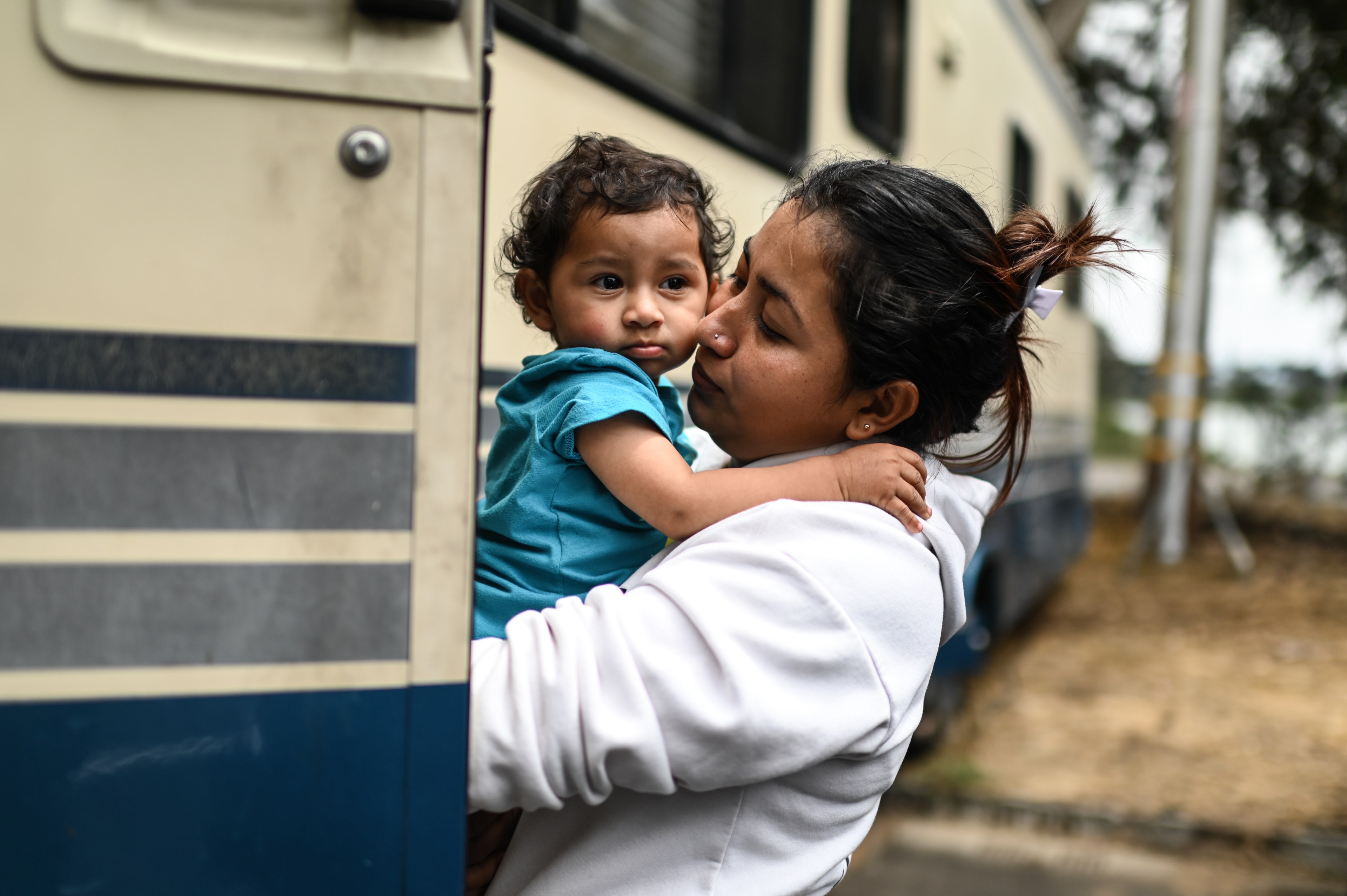 A woman wearing a white hoodie cradles a toddler dressed in blue. They stand beside a beige vehicle with blue stripes, while the toddler gazes away.