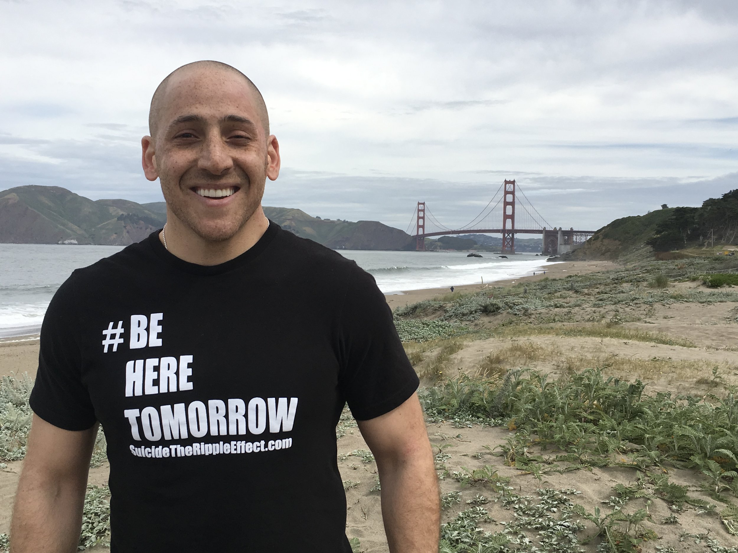 A smiling person in a black t-shirt with &quot;BE HERE TOMORROW&quot; in white text stands on a beach, with the Golden Gate Bridge and hilly landscape in the background.