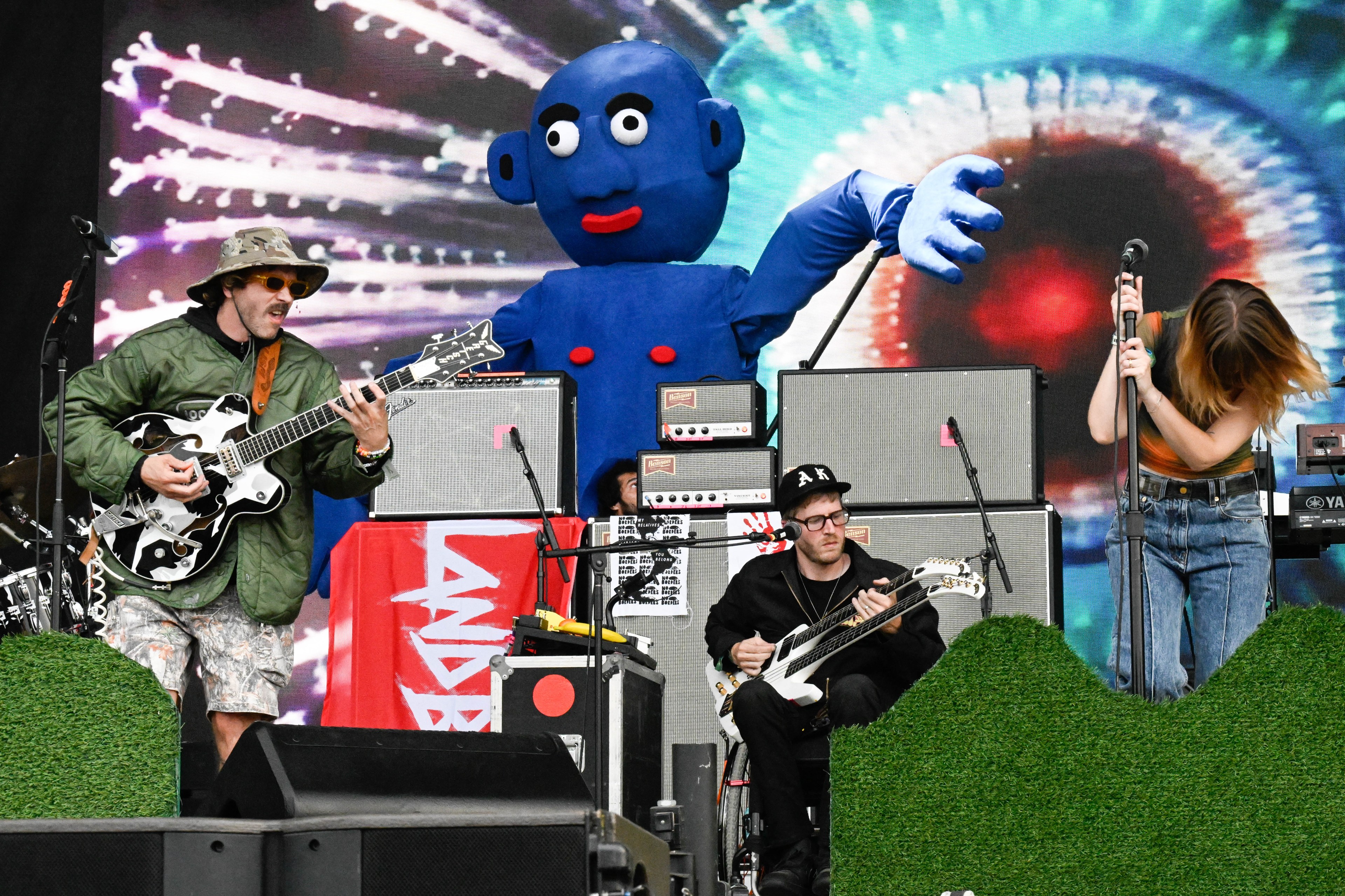 A band performs on stage with a giant blue mascot behind them, featuring a guitarist, a bassist, and a singer. The vibrant backdrop displays abstract visuals.