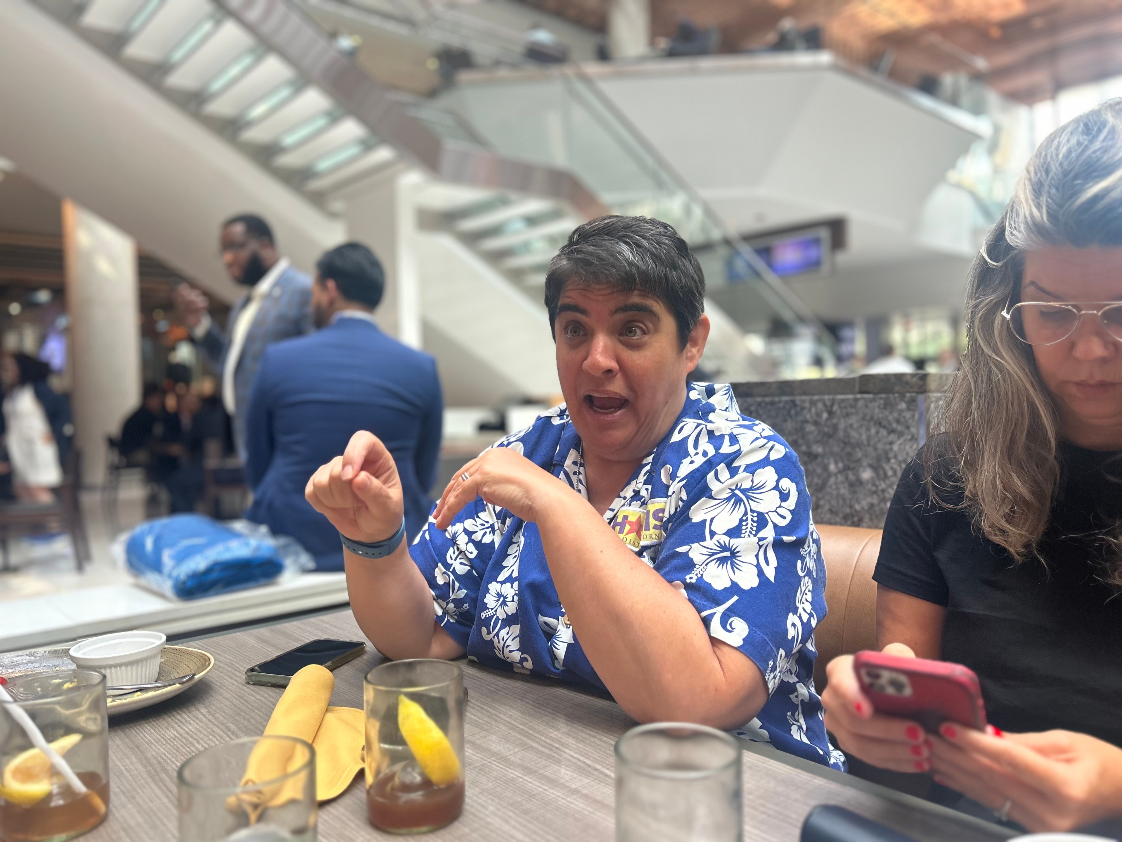 Two people are seated at a table; one is animatedly talking in a blue floral shirt, while the other, in black, is looking down at a phone.