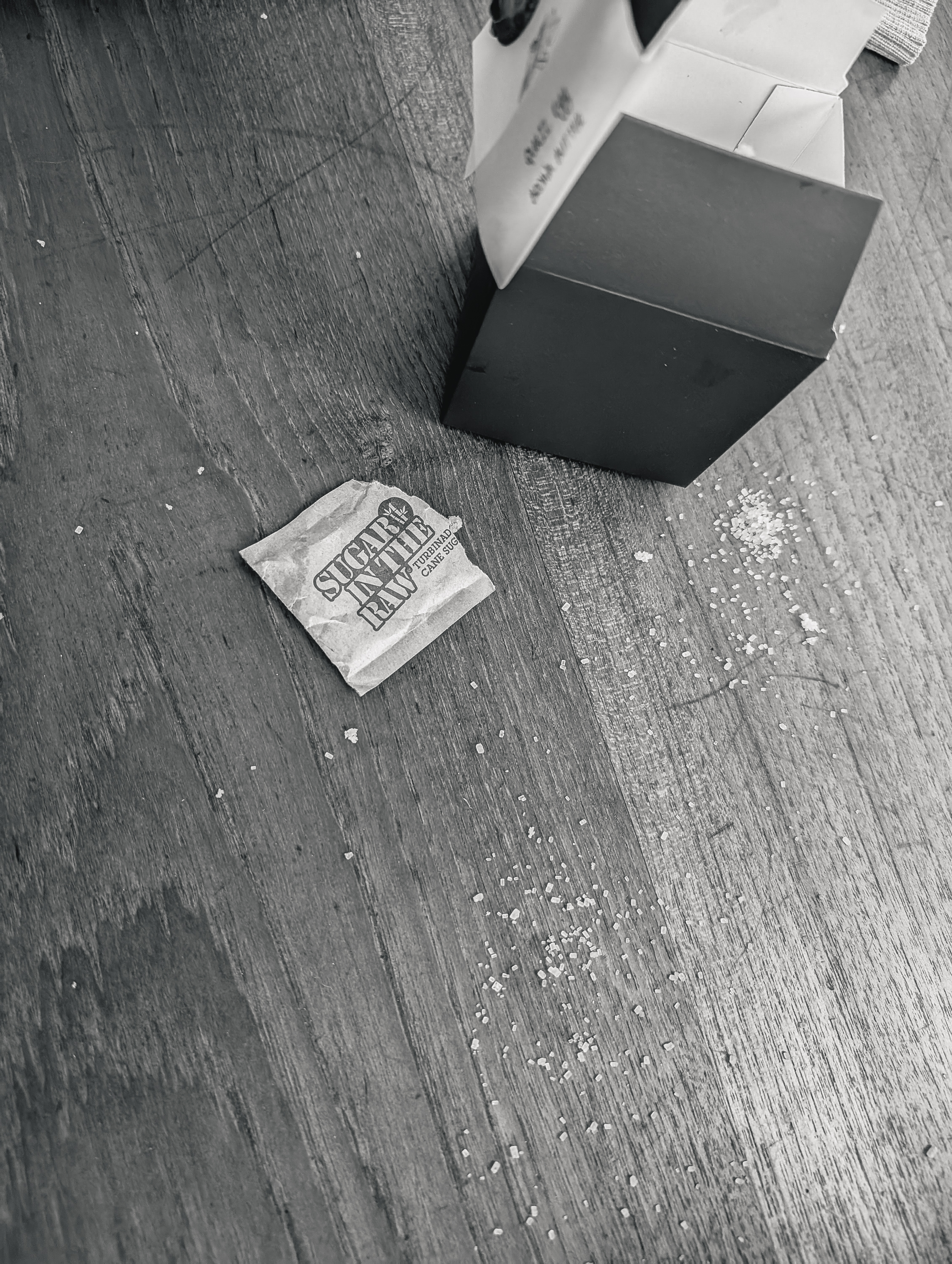 The image shows an opened black box on a wooden surface with a packet labeled &quot;Sugar In The Raw&quot; nearby and scattered sugar granules around it.