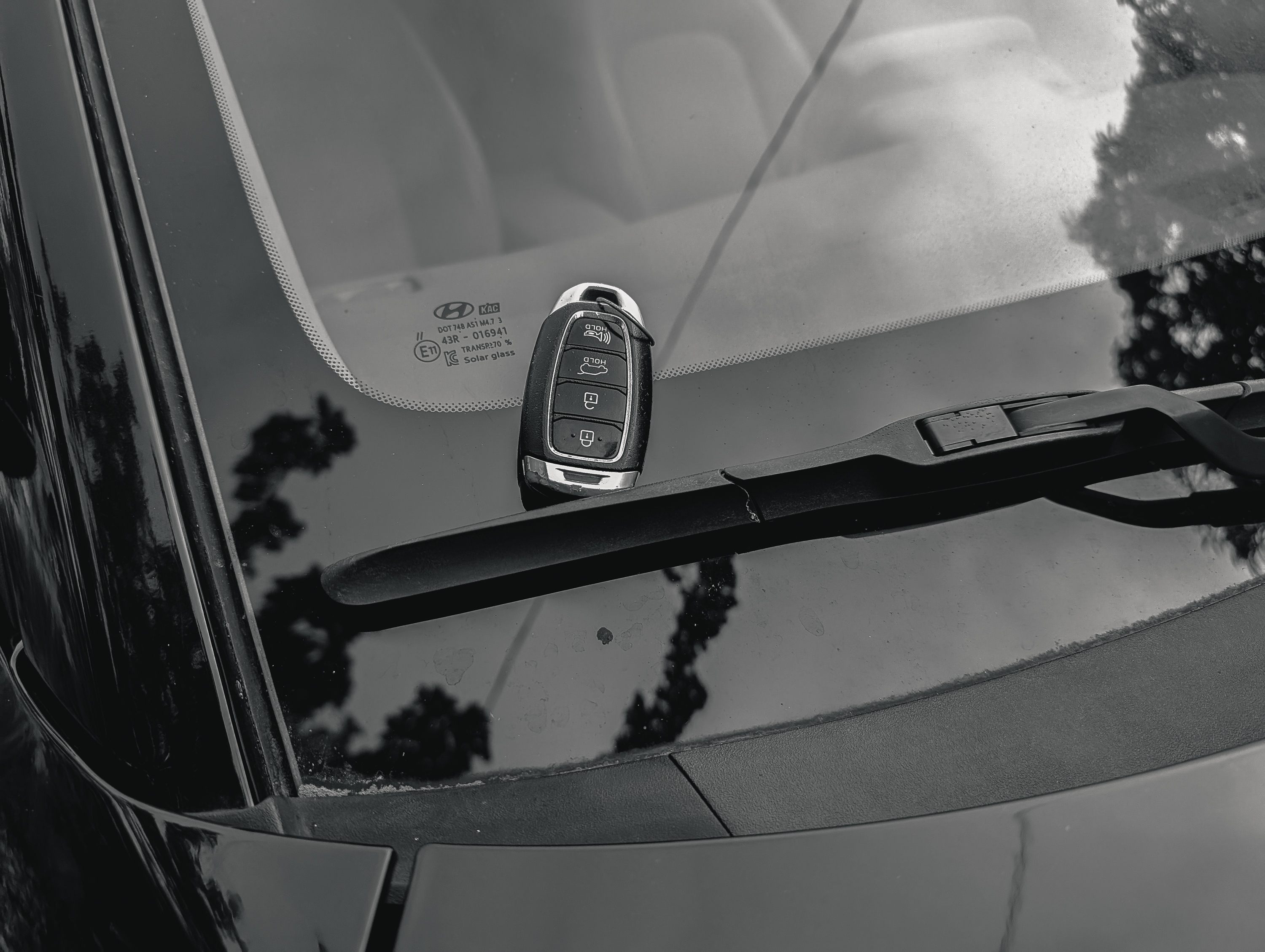 A car key fob rests on the windshield of a black Hyundai, positioned near the wiper blade. The car's interior and exterior reflections are also visible.