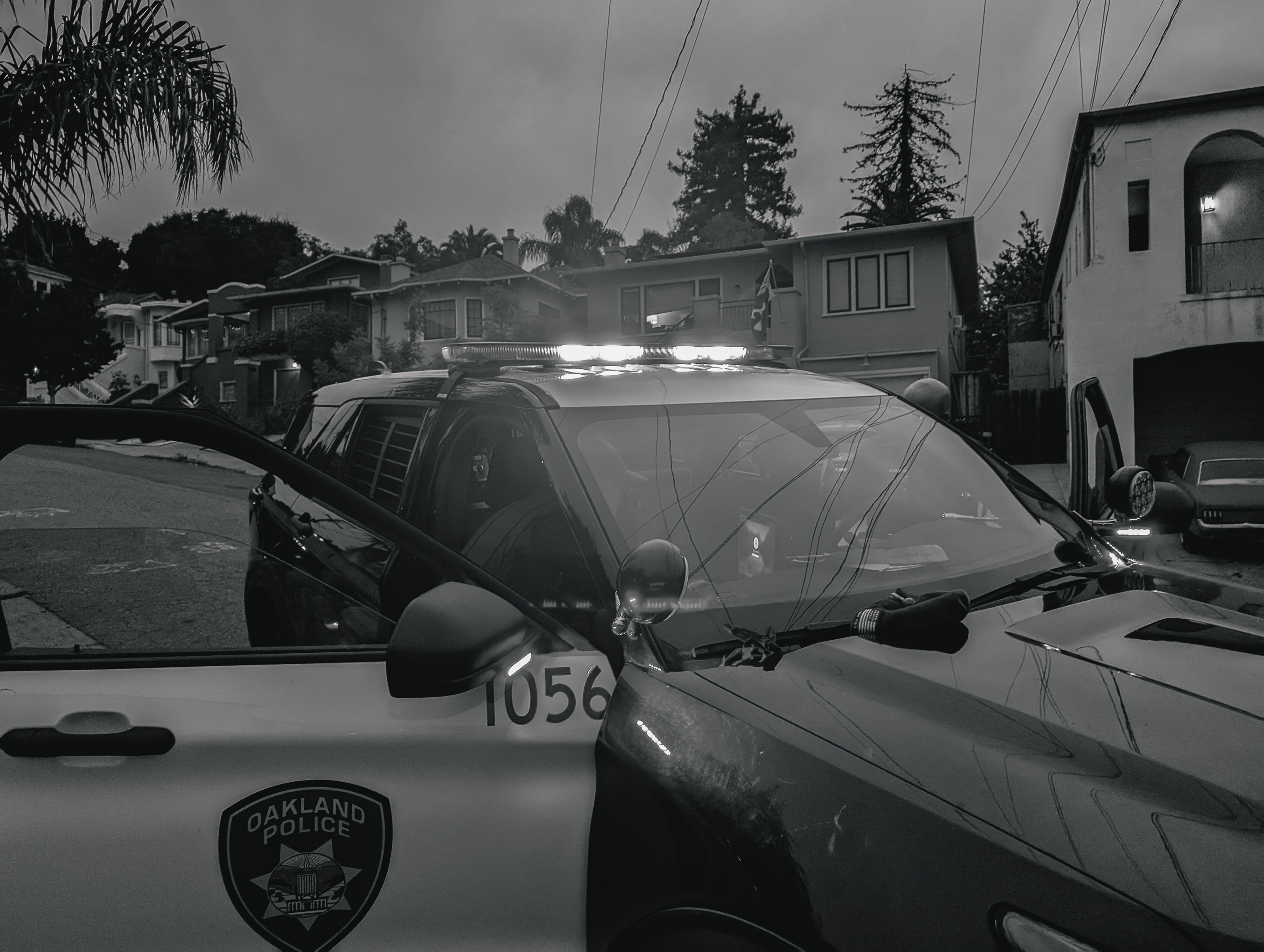 In the image, a police SUV with the Oakland Police emblem is parked on a residential street with its driver's door open and emergency lights flashing. The background shows several houses.