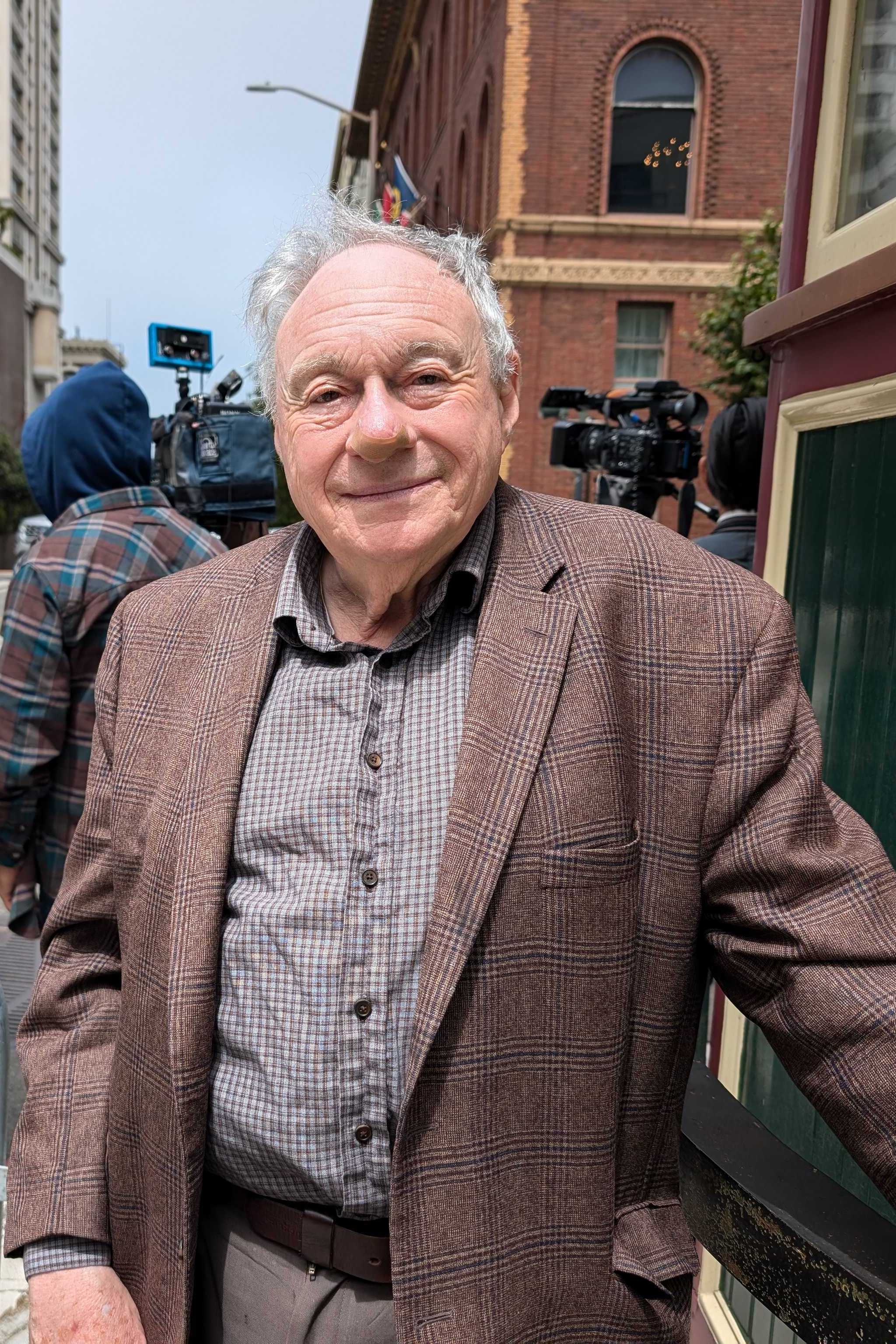 A smiling elderly man in a brown plaid jacket stands next to a building, while a group of photographers and a reporter gather behind a barricade on the street.