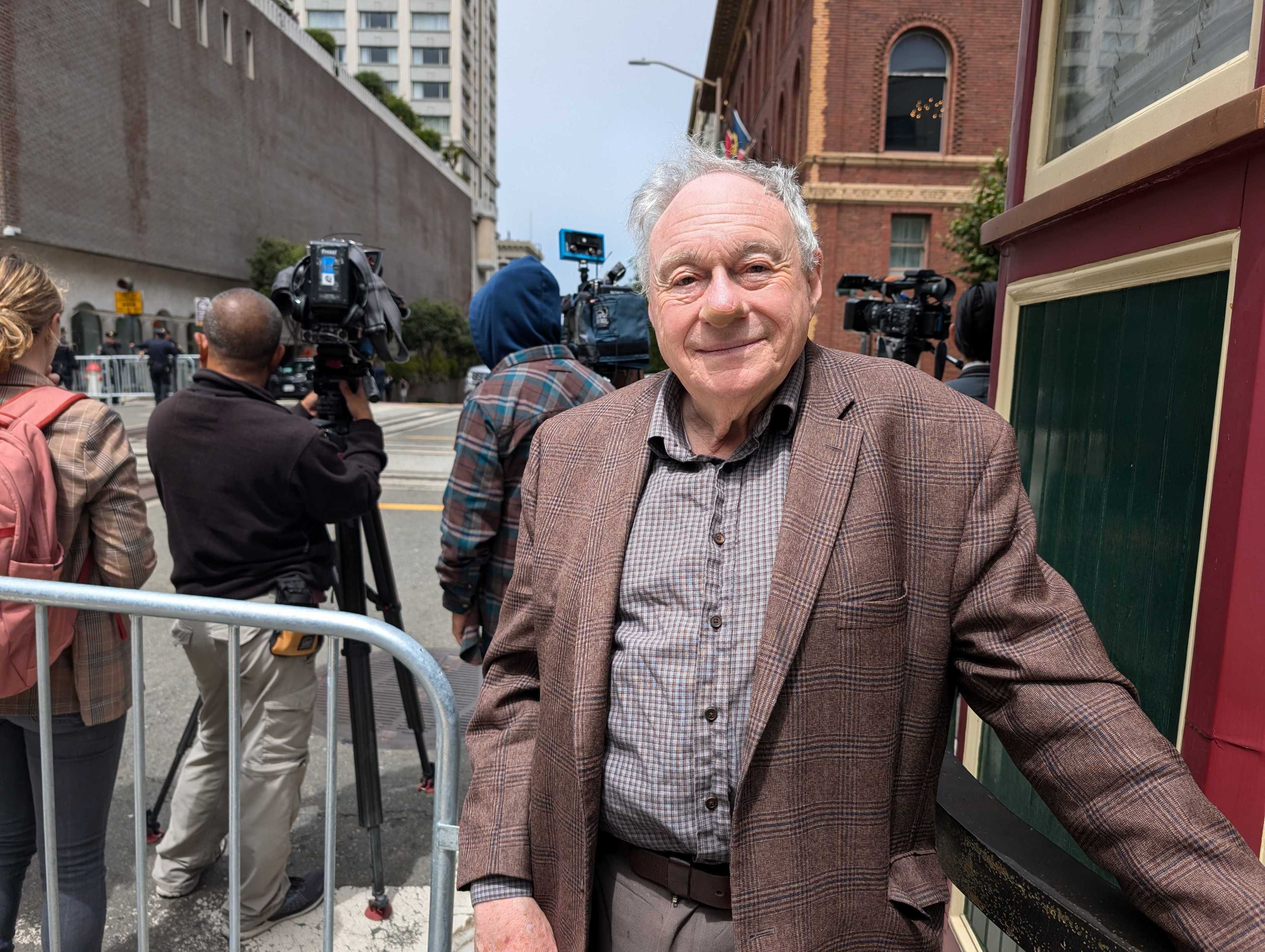 A smiling elderly man in a brown plaid jacket stands next to a building, while a group of photographers and a reporter gather behind a barricade on the street.