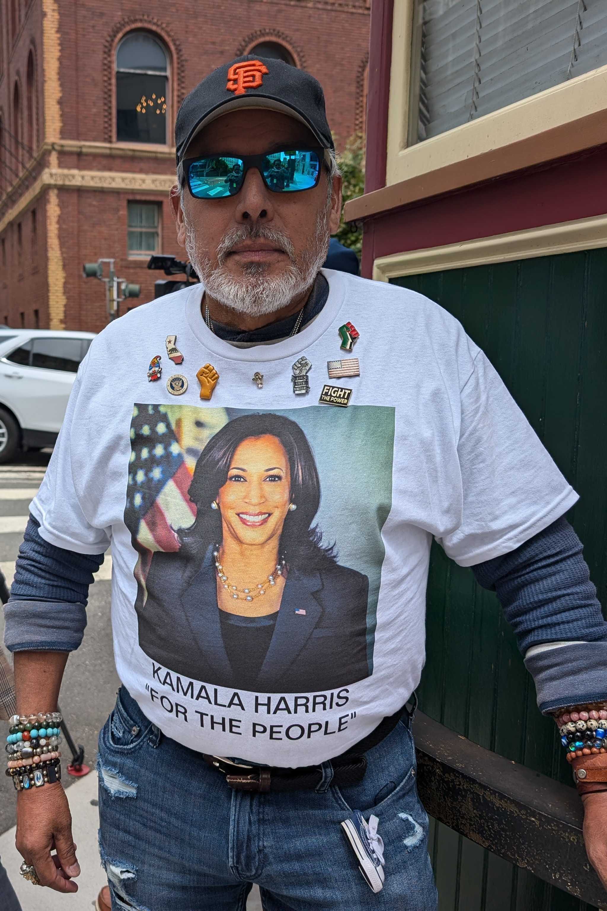 A bearded man in sunglasses wears a shirt with Kamala Harris' photo among a small crowd, including a woman in a pink backpack and videographers on a street corner.